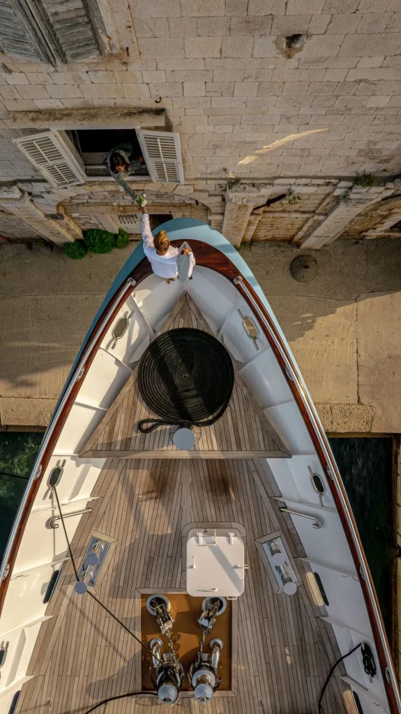A person on a docked yacht reaches up to hand a small bag to someone leaning out of a window in a stone building above, seen from an overhead perspective.