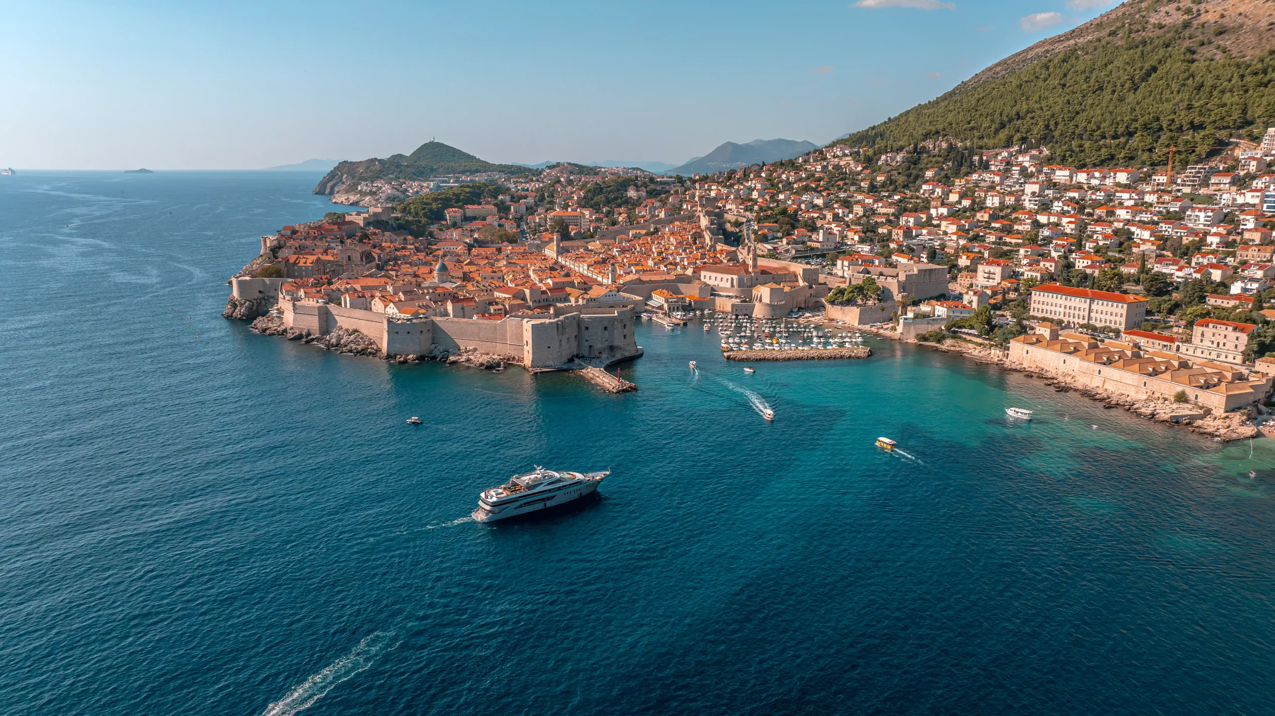 Aerial view of Dubrovnik, Croatia, showing historic stone buildings with orange roofs, the city walls, and boats sailing in the blue Adriatic Sea, with green hills in the background.