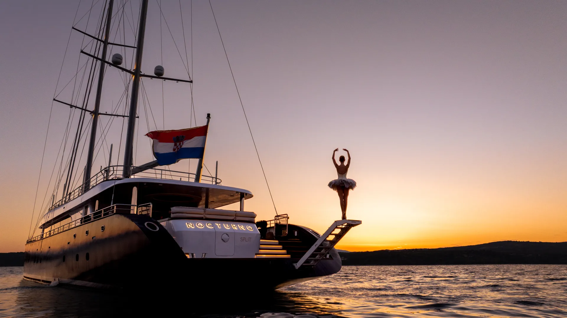 A ballerina poses on the edge of a yacht named Nocturno at sunset, with the Croatian flag flying and calm water in the foreground.
