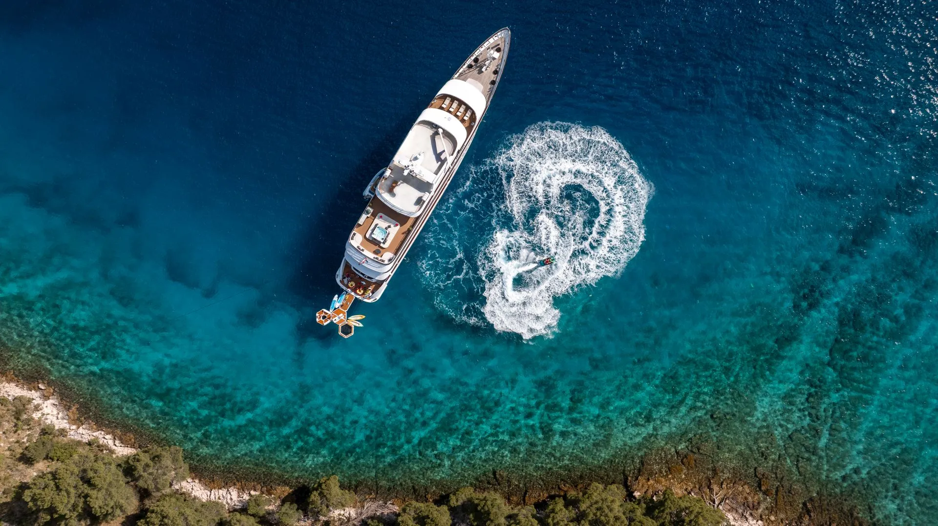 Aerial view of a yacht anchored near the shore in clear blue water, with a circular wake pattern created by a smaller boat nearby. Greenery lines the coastline at the bottom of the image.