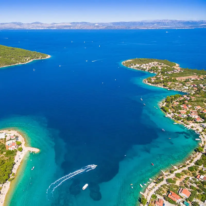 Aerial view of a coastline with clear blue water, green peninsulas, scattered houses, boats, and distant mountains under a bright blue sky.