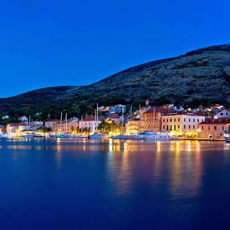 A coastal town at dusk with illuminated buildings, sailboats docked along the waterfront, and a hill rising in the background under a deep blue sky.