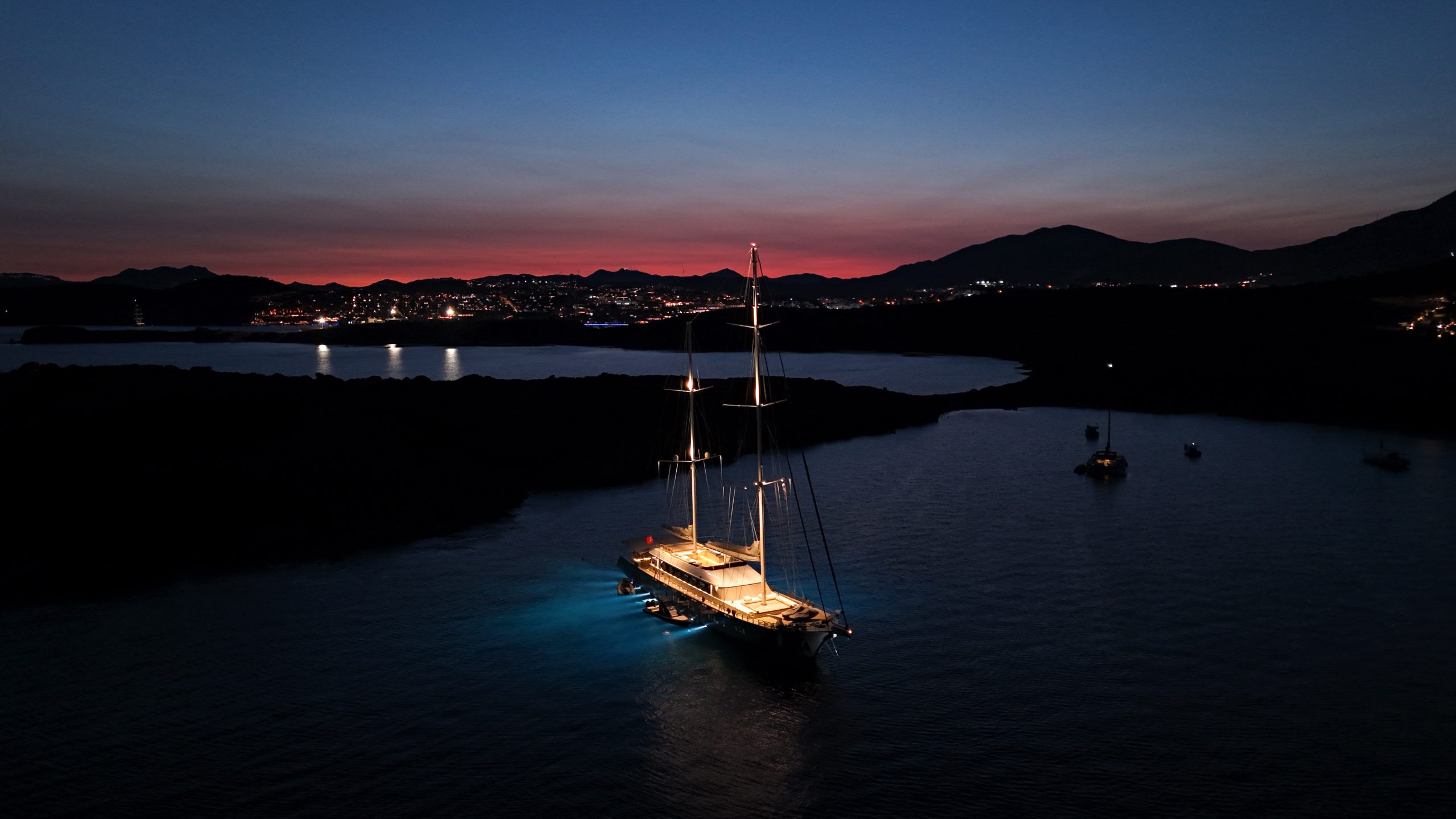 DE LOVE A sailboat with glowing lights is anchored on calm water at dusk, surrounded by a few other boats. City lights and mountains are visible in the background under a darkening sky.