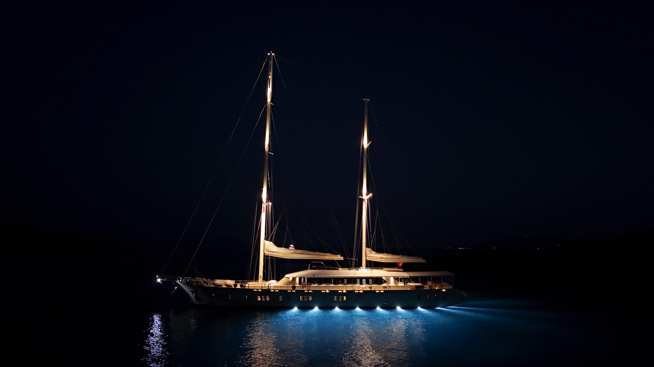 DE LOVE A large sailboat with illuminated masts and deck lights floats on calm, dark water at night, casting a soft glow on the surrounding water.