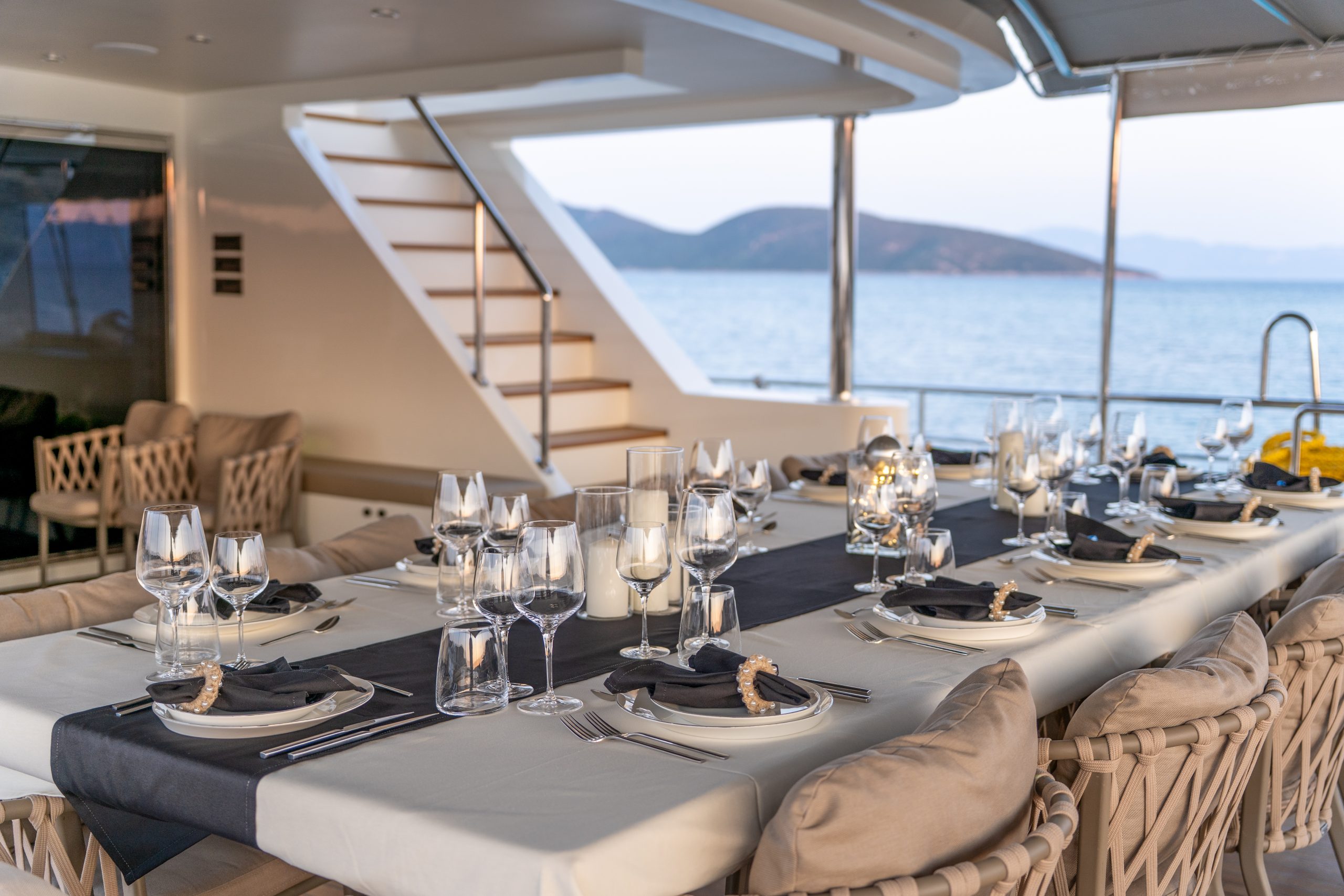 DE LOVE An elegant outdoor dining table is set with glassware, plates, and candles on a boat deck, overlooking calm water and distant mountains at sunset. Cushioned chairs surround the table, creating a luxurious atmosphere.