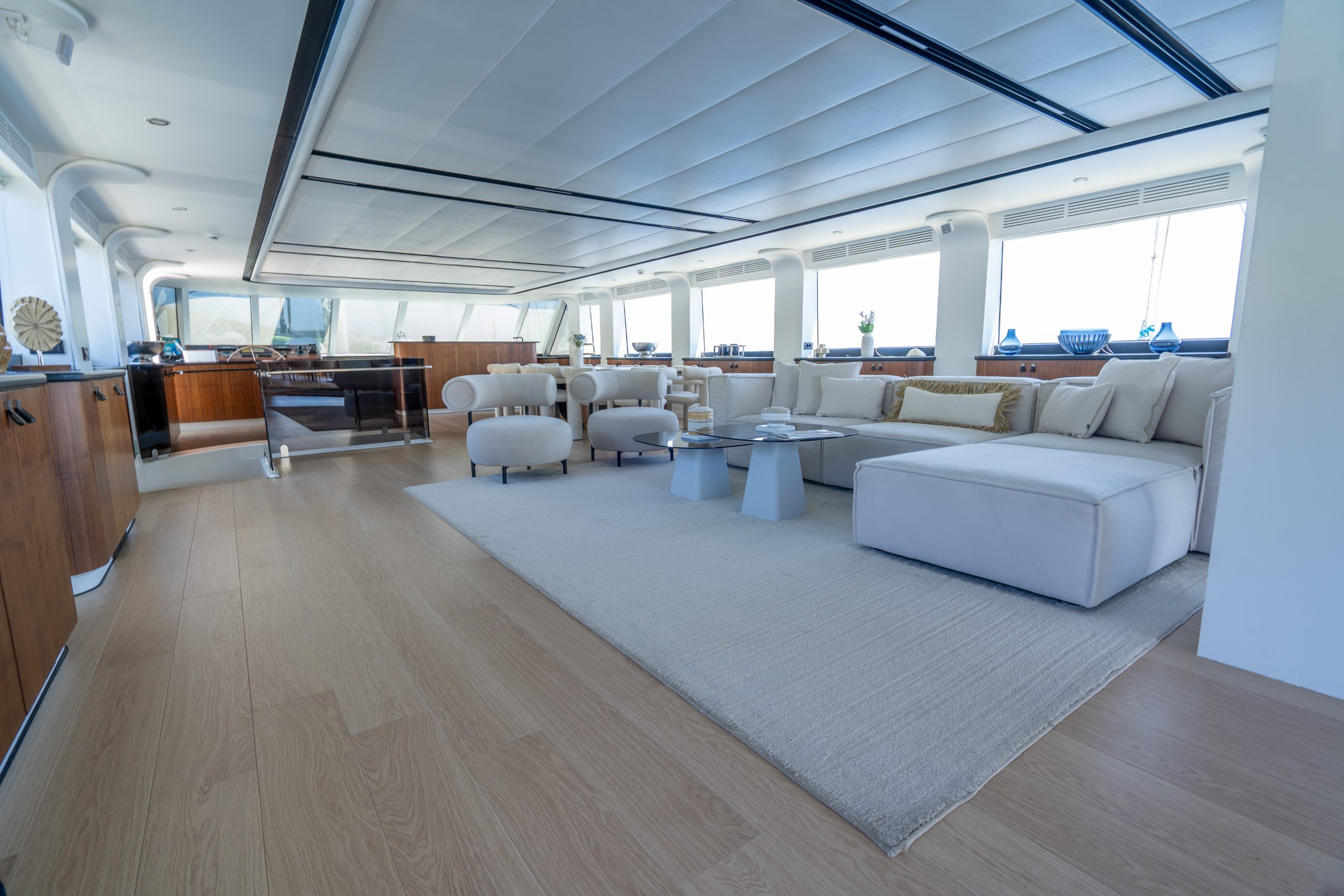DE LOVE Spacious, modern yacht interior with light wood floors, a large beige sectional sofa, round armchairs, low tables, and panoramic windows letting in natural light. The design features neutral tones and minimalist decor.