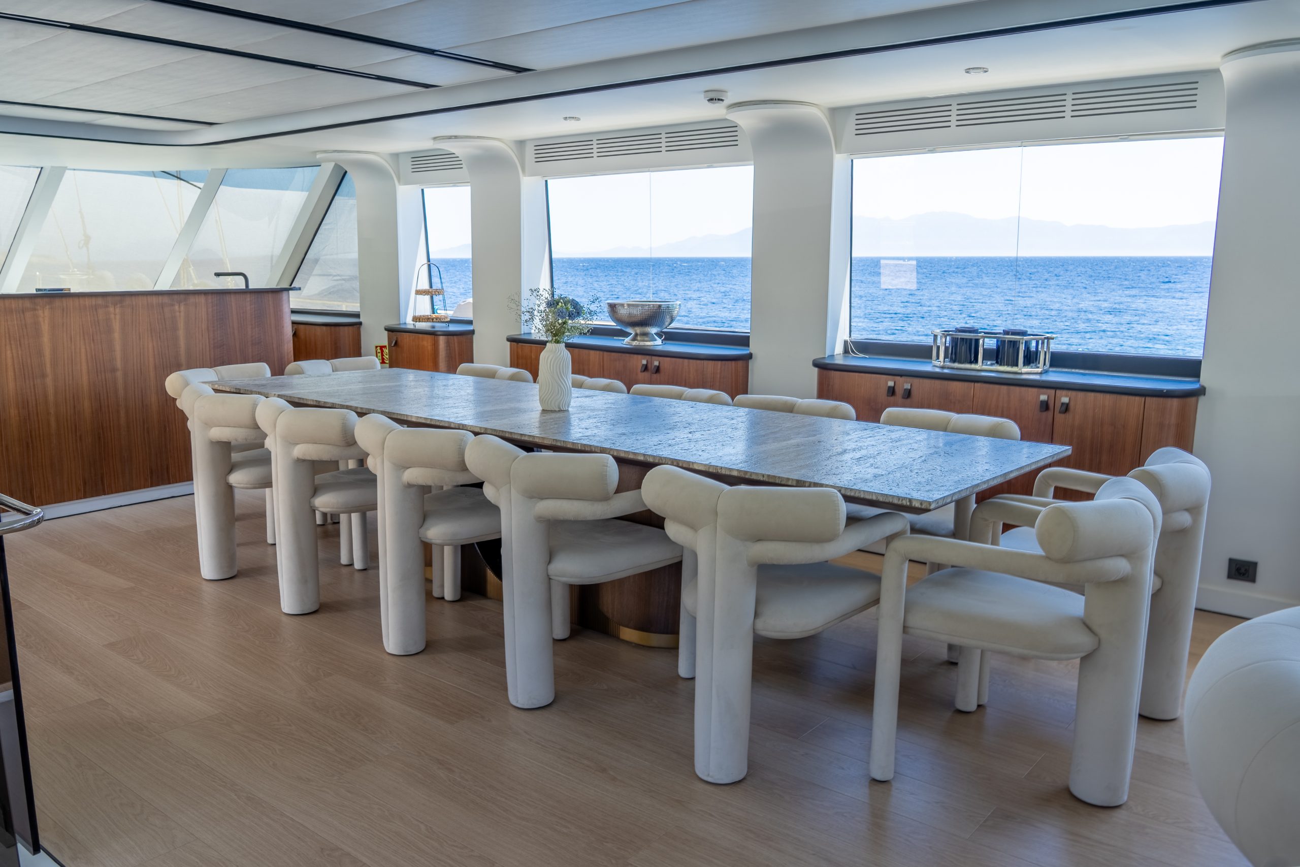 DE LOVE A modern dining area on a yacht with a long wooden table, white cushioned chairs, large windows overlooking the ocean, and light wood flooring.