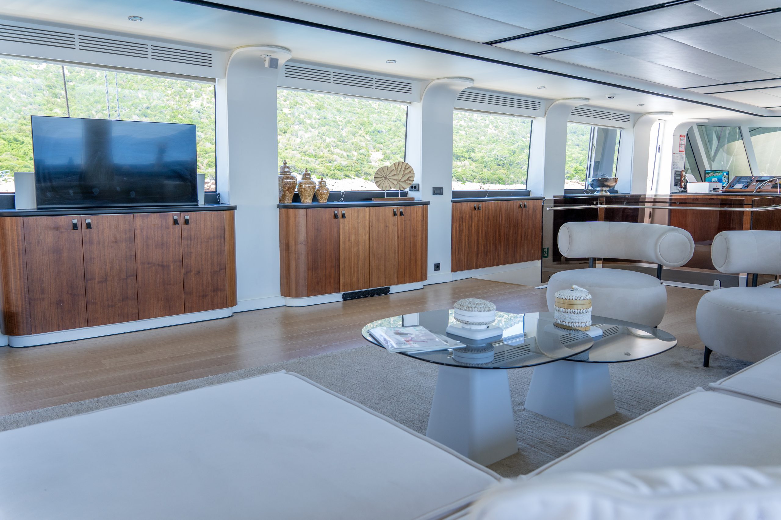 DE LOVE Modern yacht interior with large windows, wood cabinets, cream-colored chairs and sofa, a glass coffee table with decor, and a flat-screen TV. Natural light fills the room, highlighting the bright and inviting atmosphere.