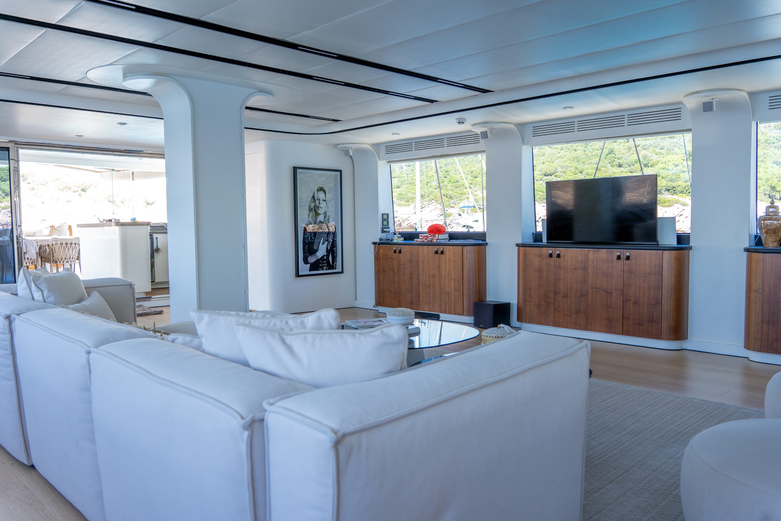 DE LOVE A modern yacht lounge with white sofas, a glass coffee table, wooden cabinets, a large flat-screen TV, and large windows overlooking water and greenery. A piece of art hangs on the wall near the sliding glass door.