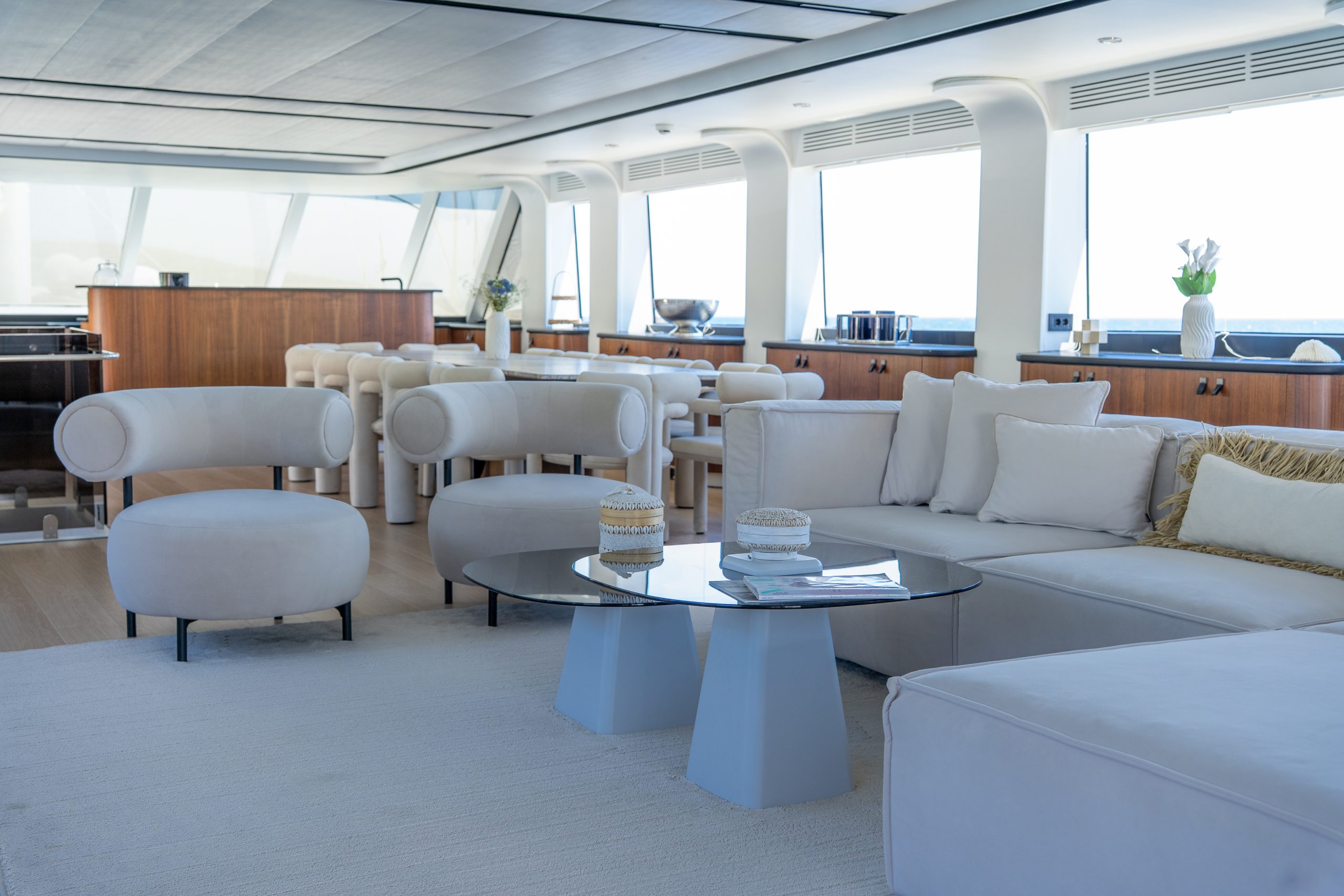 DE LOVE A modern yacht interior with white cushioned seating, round chairs, a glass coffee table, dining area, and large windows letting in natural light. The space is decorated in neutral tones with some wooden accents.