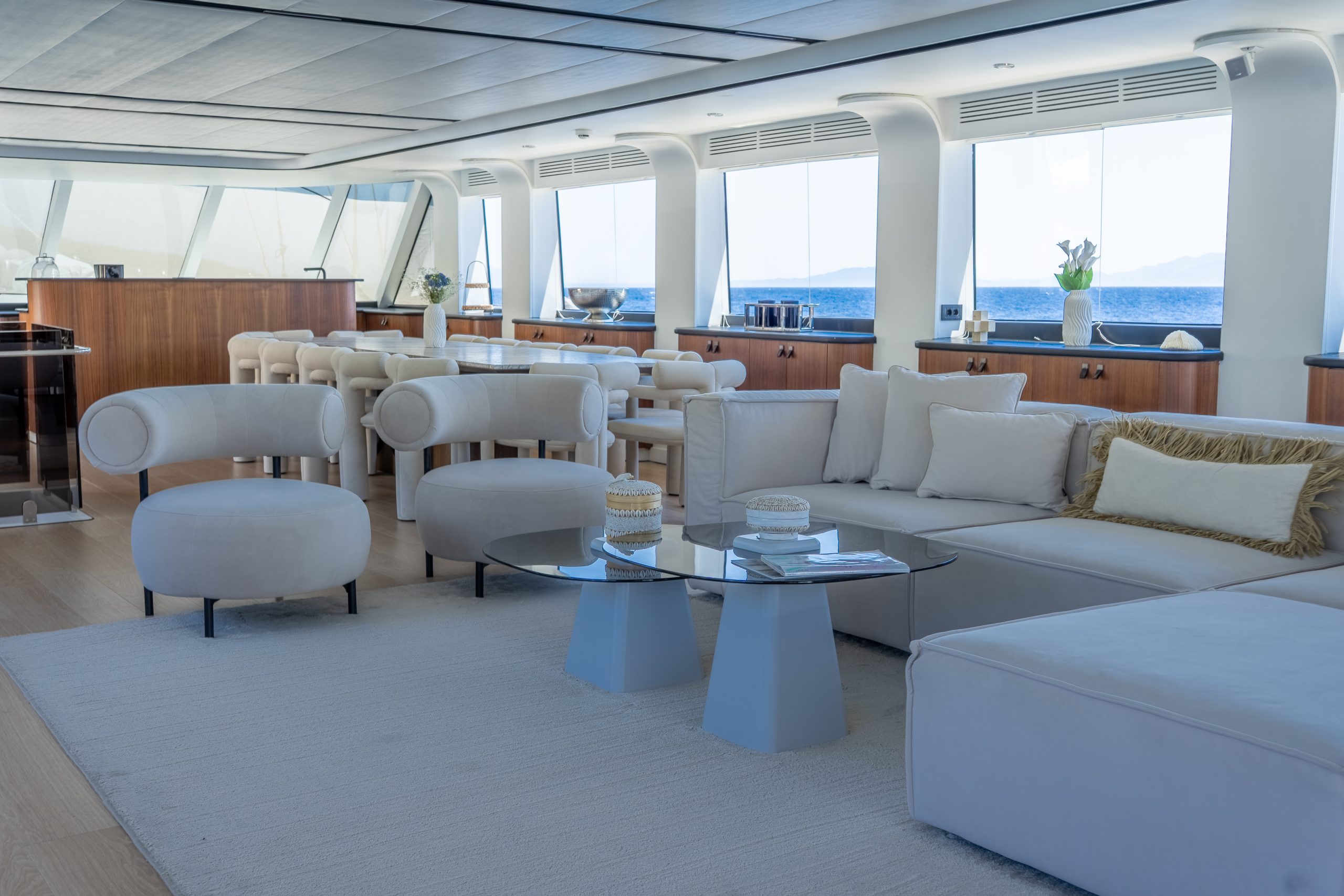 DE LOVE Modern yacht interior with large windows, cream-colored sofas and chairs, two round glass coffee tables, and a dining area in the background. Natural light fills the space, offering ocean and mountain views.