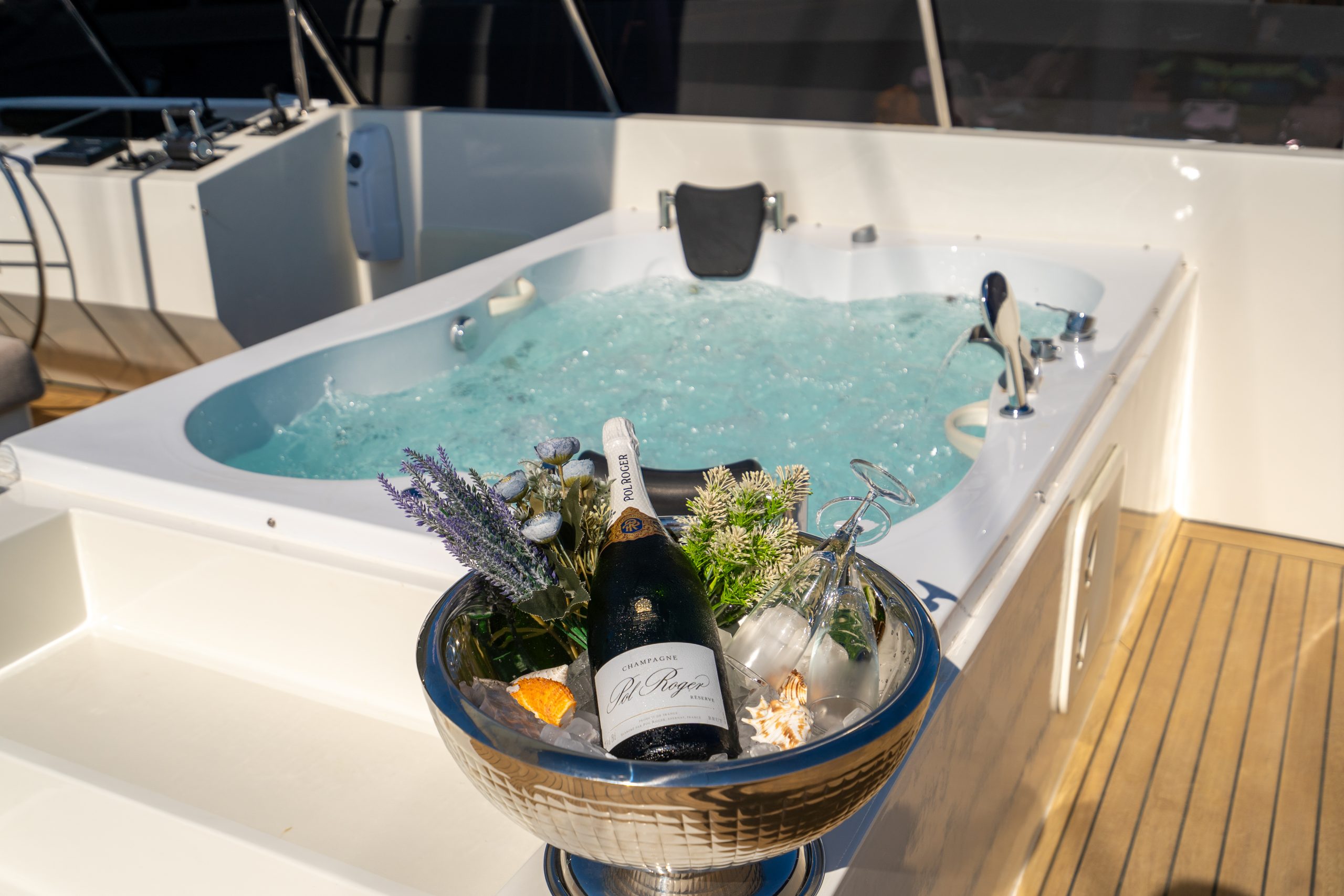 DE LOVE A bottle of champagne sits chilled in a bucket with flowers and glasses beside a bubbling jacuzzi on the deck of a luxury boat under bright sunlight.