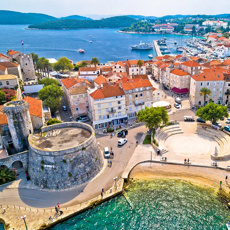 Aerial view of a coastal town with historic stone buildings, red-tiled roofs, a round fort, a small marina, boats on blue water, and lush green hills in the background.
