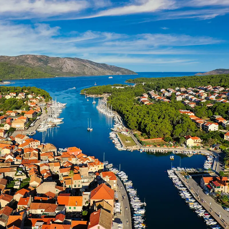 Aerial view of a coastal town with red-roofed buildings, marinas filled with boats, a narrow waterway, green trees, and mountains in the background under a blue sky.