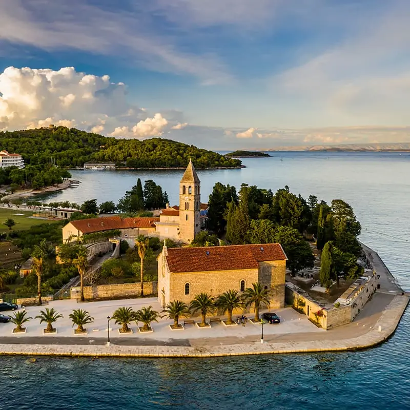 A coastal village with red-roofed stone buildings, a church with a tall bell tower, palm trees, and calm blue water, surrounded by greenery under a partly cloudy sky.