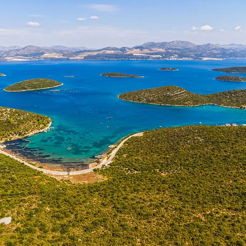 Aerial view of a blue bay with several small green islands, surrounded by lush green hills and a few buildings along the shoreline, under a mostly clear sky.