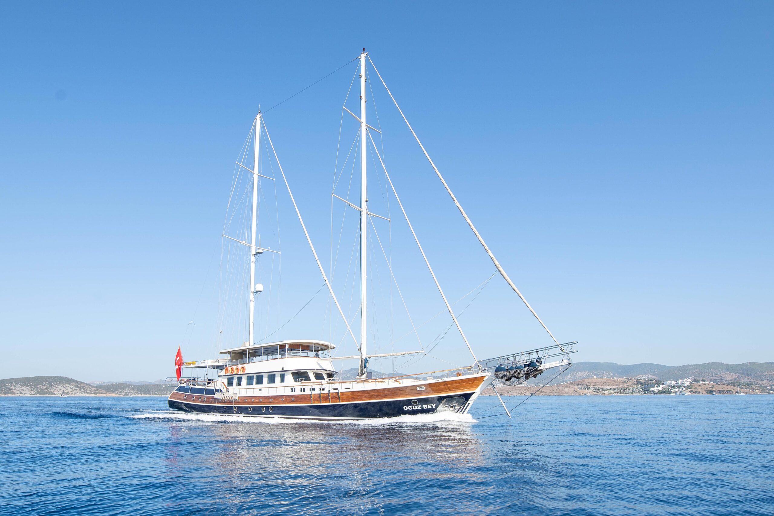 OGUZ BEY A large, elegant sailing yacht with two masts cruises on calm blue water under a clear sky, with distant hills visible in the background.