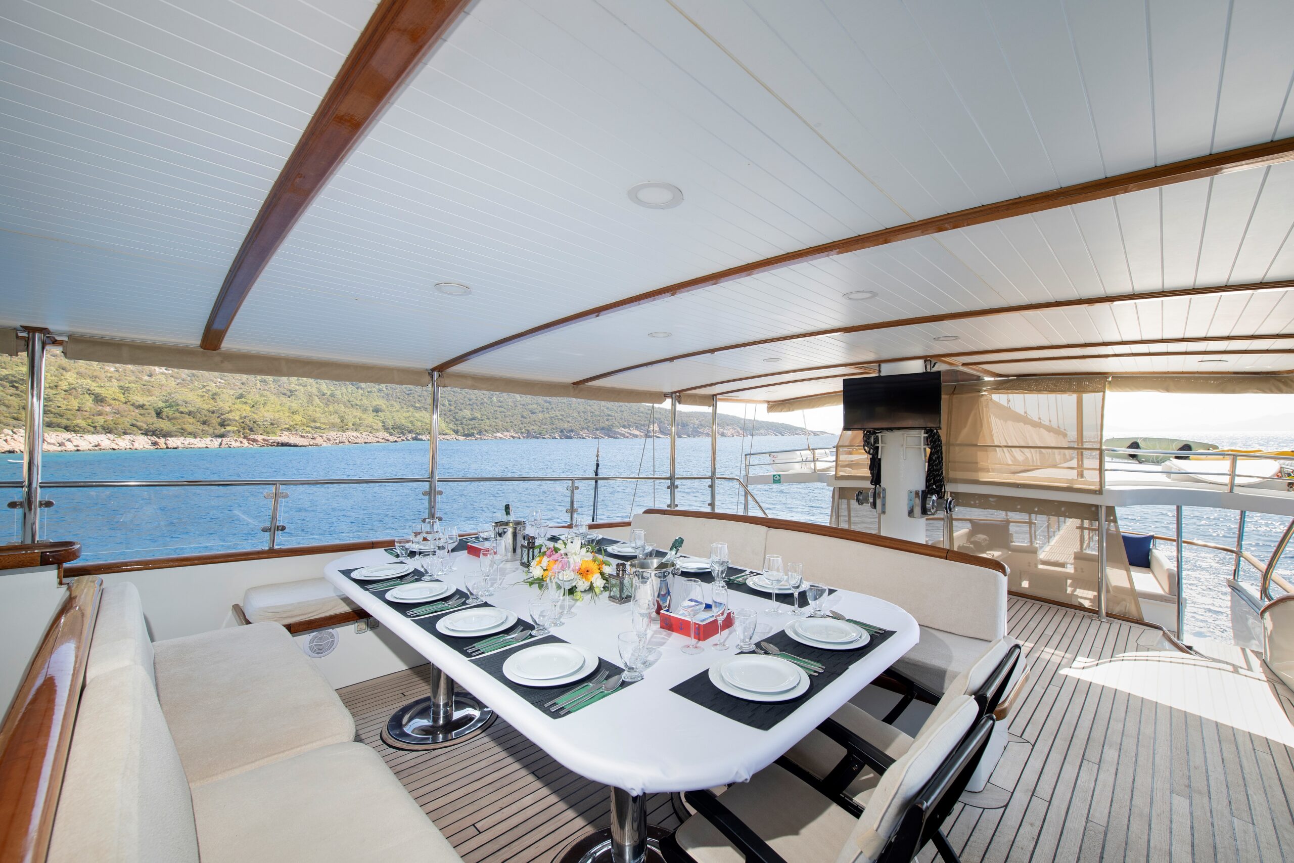 OGUZ BEY Elegant dining table set for a meal on the deck of a yacht, overlooking calm blue water and a forested coastline under a shaded canopy.