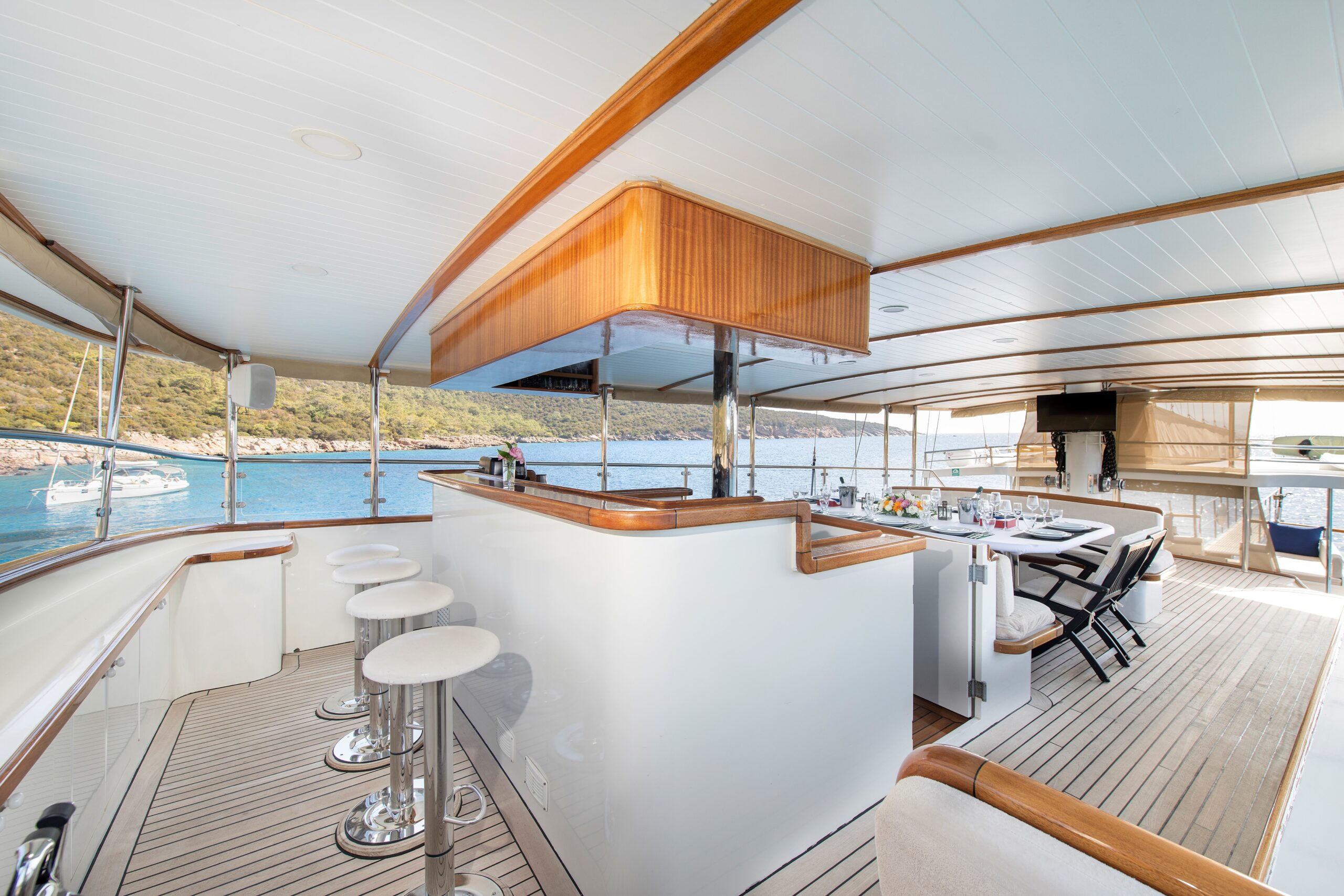 OGUZ BEY A spacious yacht deck featuring a bar with stools, a dining table set for a meal, and scenic views of the sea and nearby coastline through large open windows.