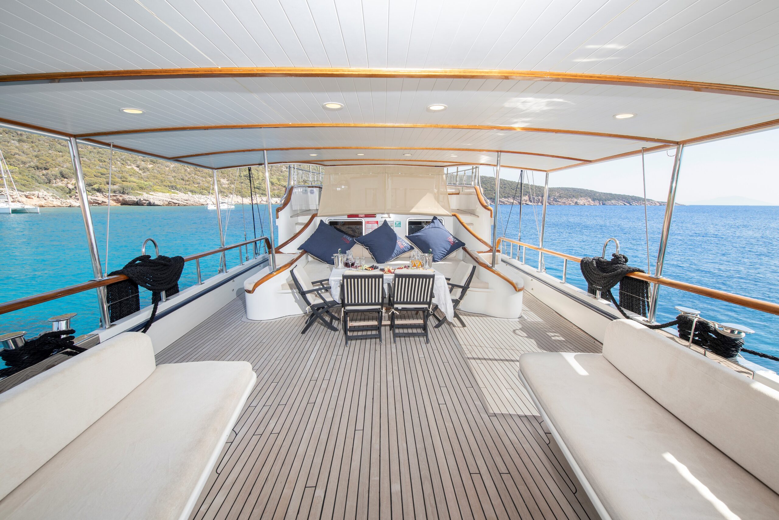 OGUZ BEY The image shows the deck of a luxury yacht with cushioned seating, a dining table set with chairs and pillows, overlooking clear blue water and a coastline under a bright sky.
