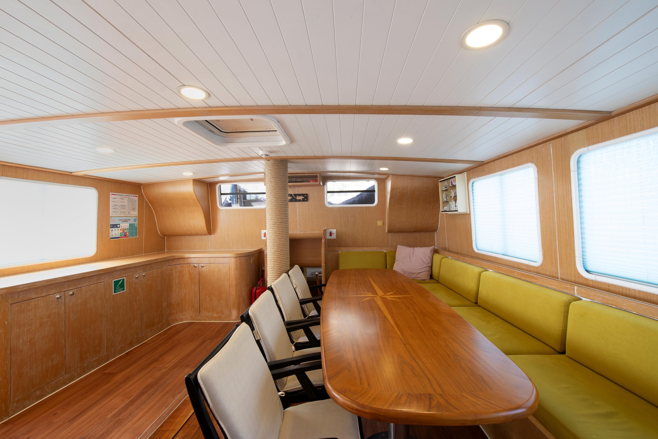 OGUZ BEY A conference room with wood-paneled walls and ceiling, a long wooden table, cushioned green benches on one side, white office chairs on the other, and large windows letting in natural light.