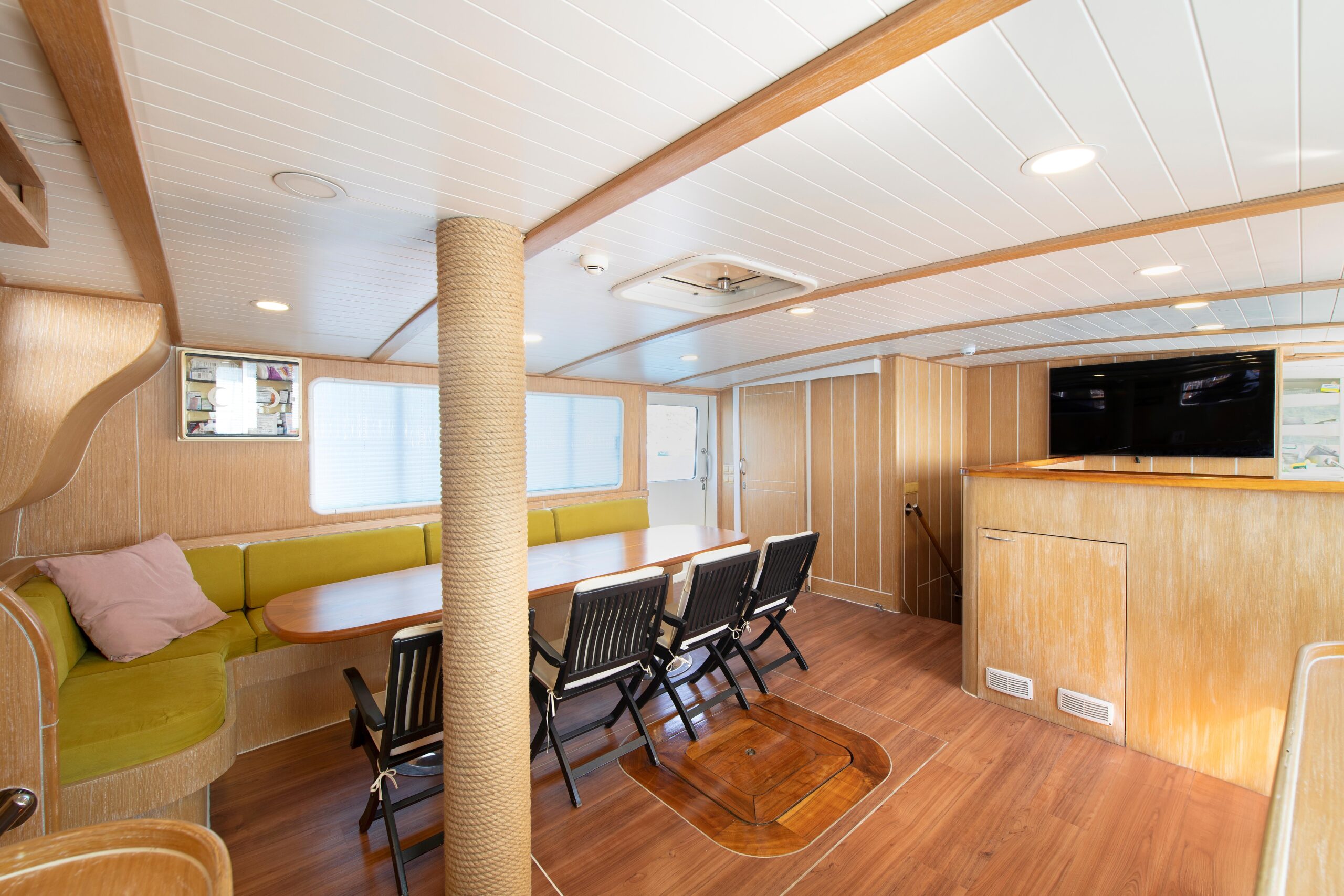 OGUZ BEY A bright, modern boat interior with wood paneling, a green cushioned bench, a wooden table with six chairs, a large TV mounted on the wall, and natural light coming through the windows.