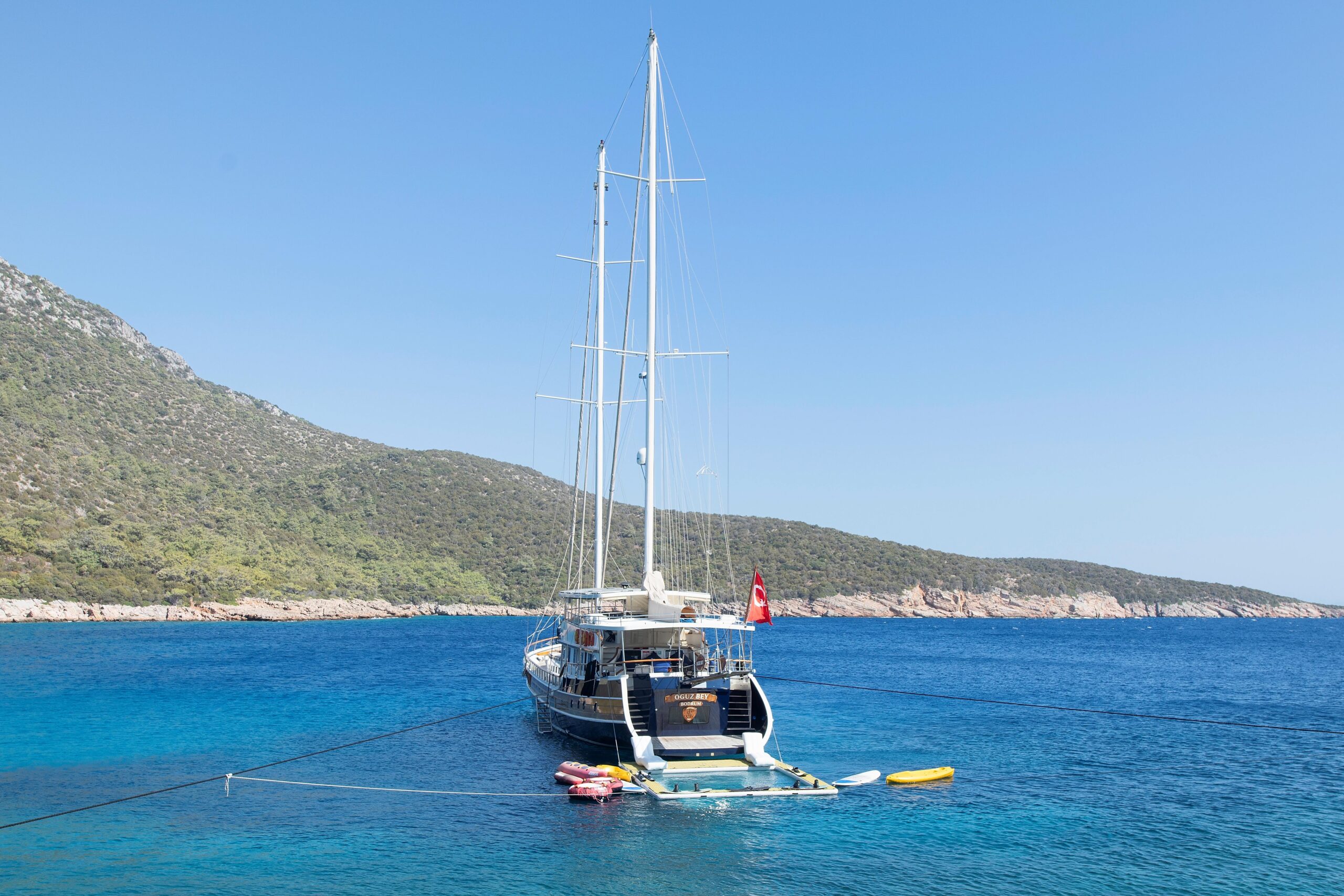 OGUZ BEY A large sailboat is anchored in clear blue water near a hilly, green coastline. Several small watercraft are tied behind the boat, and a Turkish flag is visible at the stern. The sky is clear and sunny.