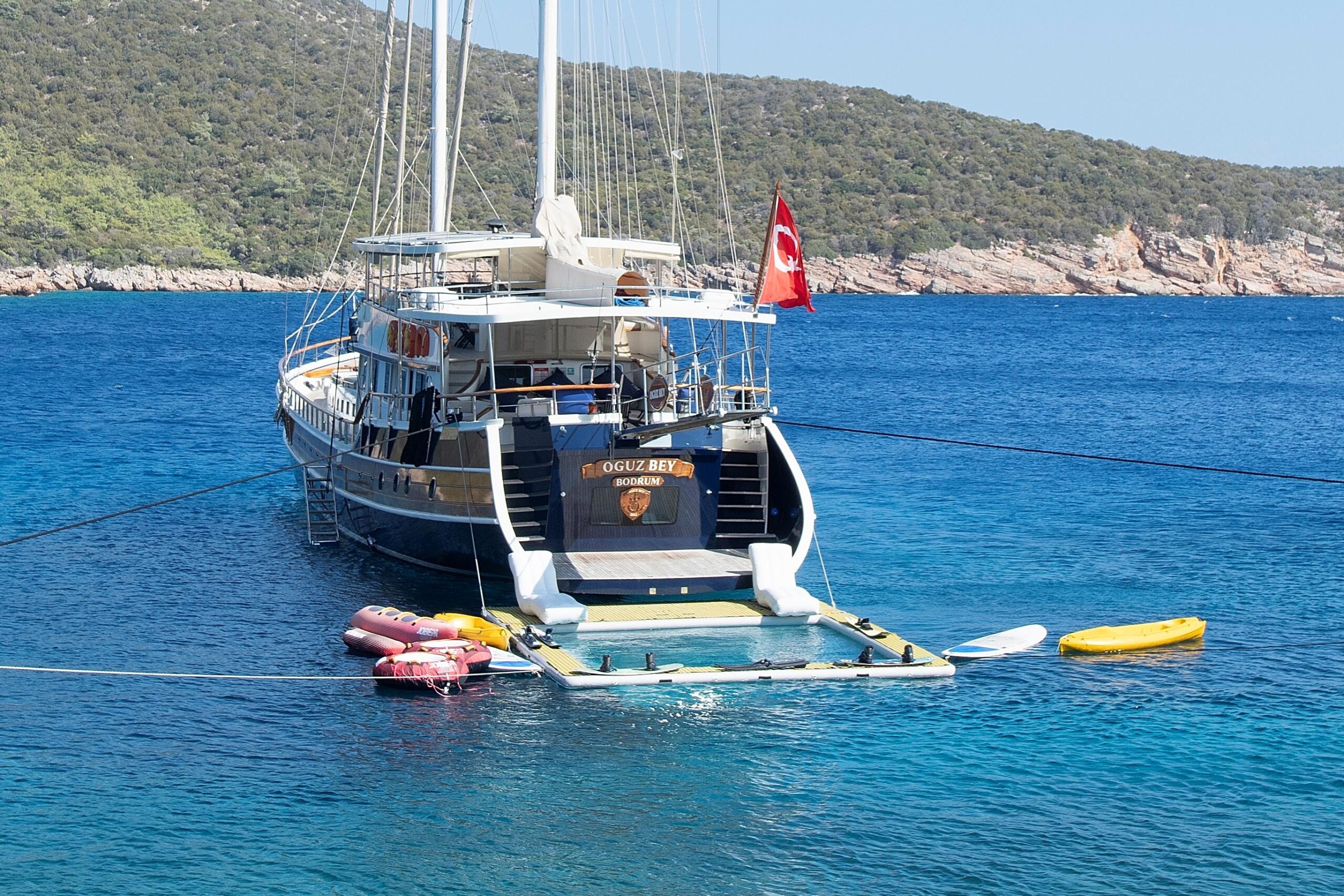 OGUZ BEY A large yacht with a Turkish flag is anchored in clear blue water near a green, rocky shoreline. Several colorful kayaks and paddleboards float nearby, secured to the boat.