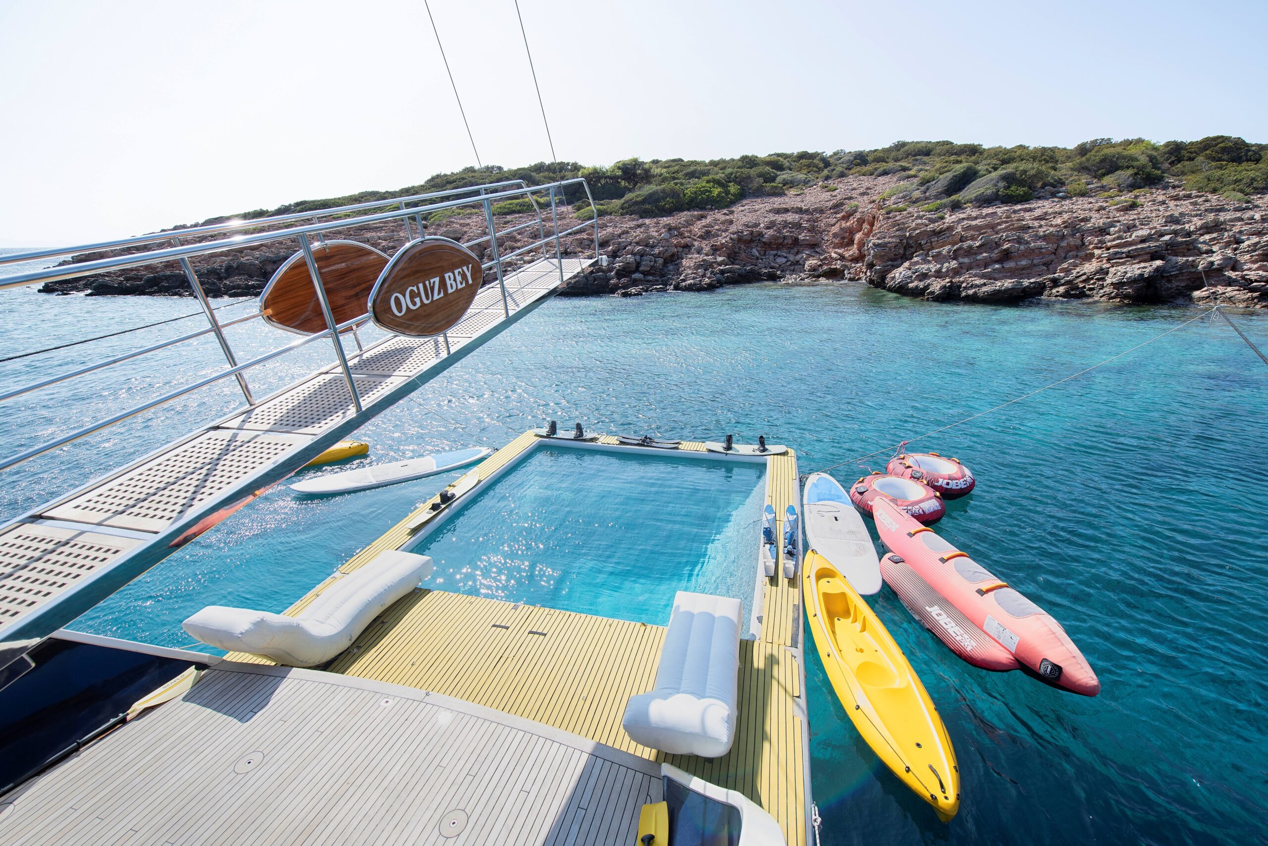 OGUZ BEY A luxury yacht with a built-in pool is anchored in clear blue water near a rocky coastline. Paddleboards, kayaks, and inflatables float nearby, and the yachts name OGUZ BEY is shown on a sign.