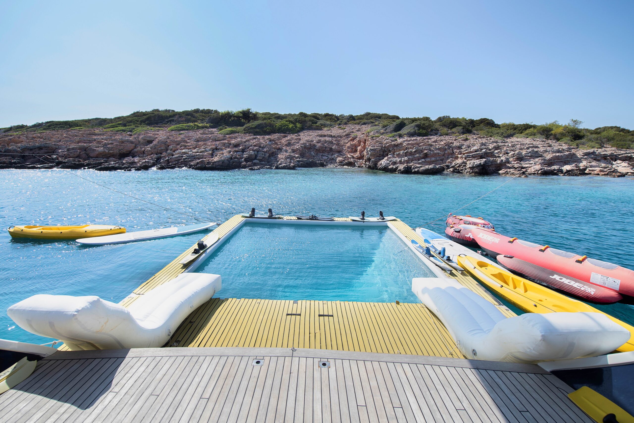 OGUZ BEY A luxurious yacht deck features a rectangular pool overlooking clear blue water, with inflatable loungers on the deck and several yellow and red kayaks and paddleboards floating nearby. Rocky, green shoreline in the background.