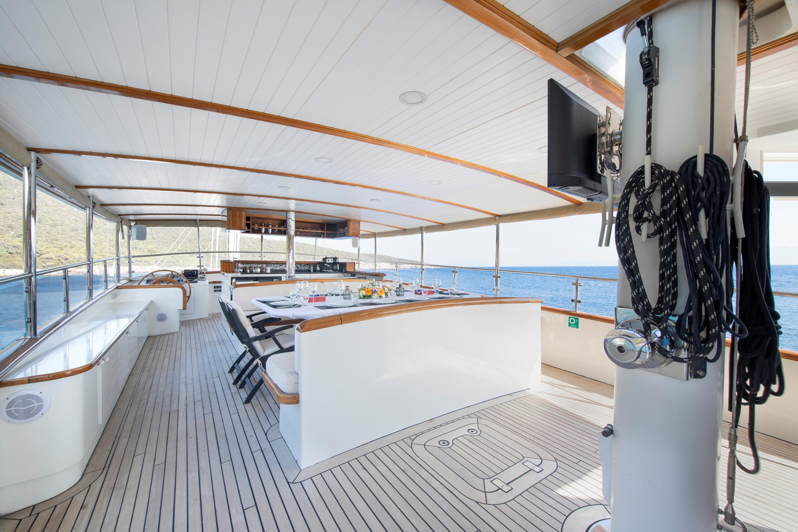 OGUZ BEY A spacious yacht deck with a dining area set for a meal, white seating, wood accents, and a view of the sea and coastline in the background under a covered roof. Nautical equipment hangs on the right side.