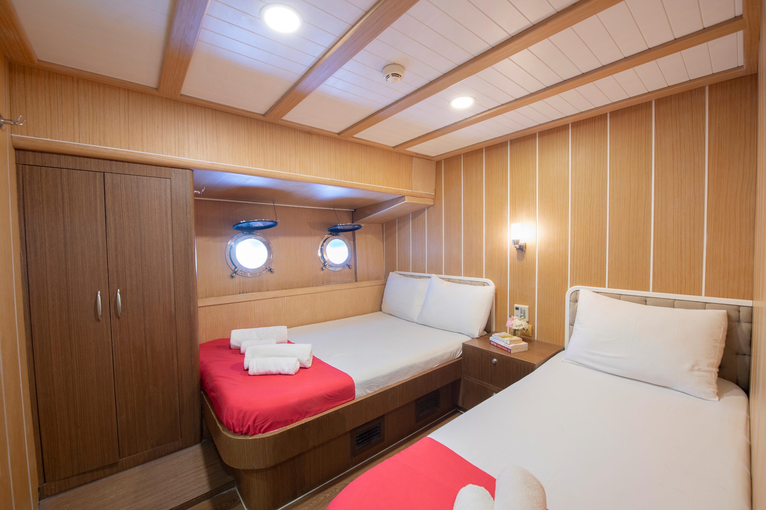 OGUZ BEY A cozy ship cabin with two beds, each with white sheets and red runners. Rolled towels sit on the beds. Wooden walls, a wardrobe, and two round porthole windows complete the warm, inviting space.