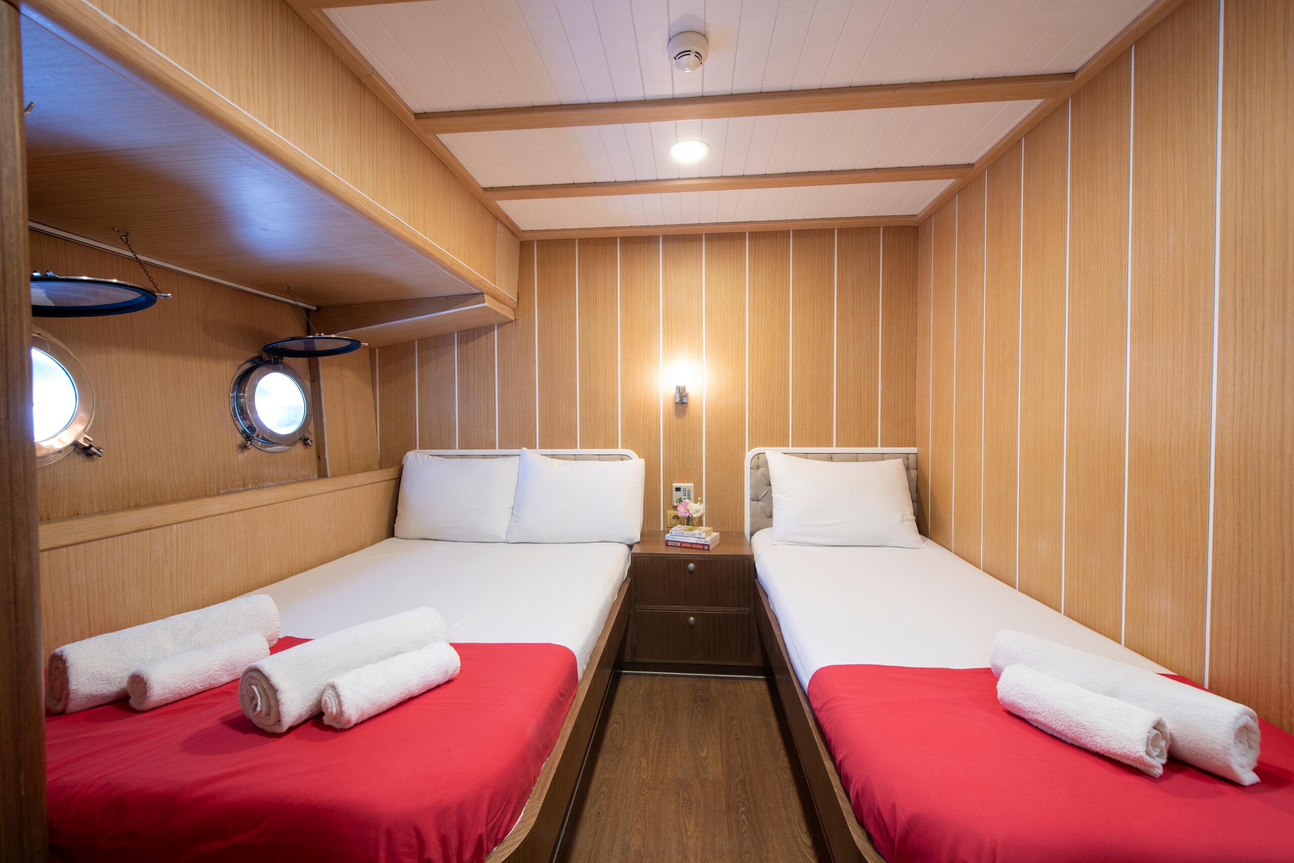 OGUZ BEY A cozy cabin with two single beds, each with white pillows, red blankets, and rolled white towels. Wooden walls and ceiling, two round porthole windows, a reading light, and a small bedside table are visible.