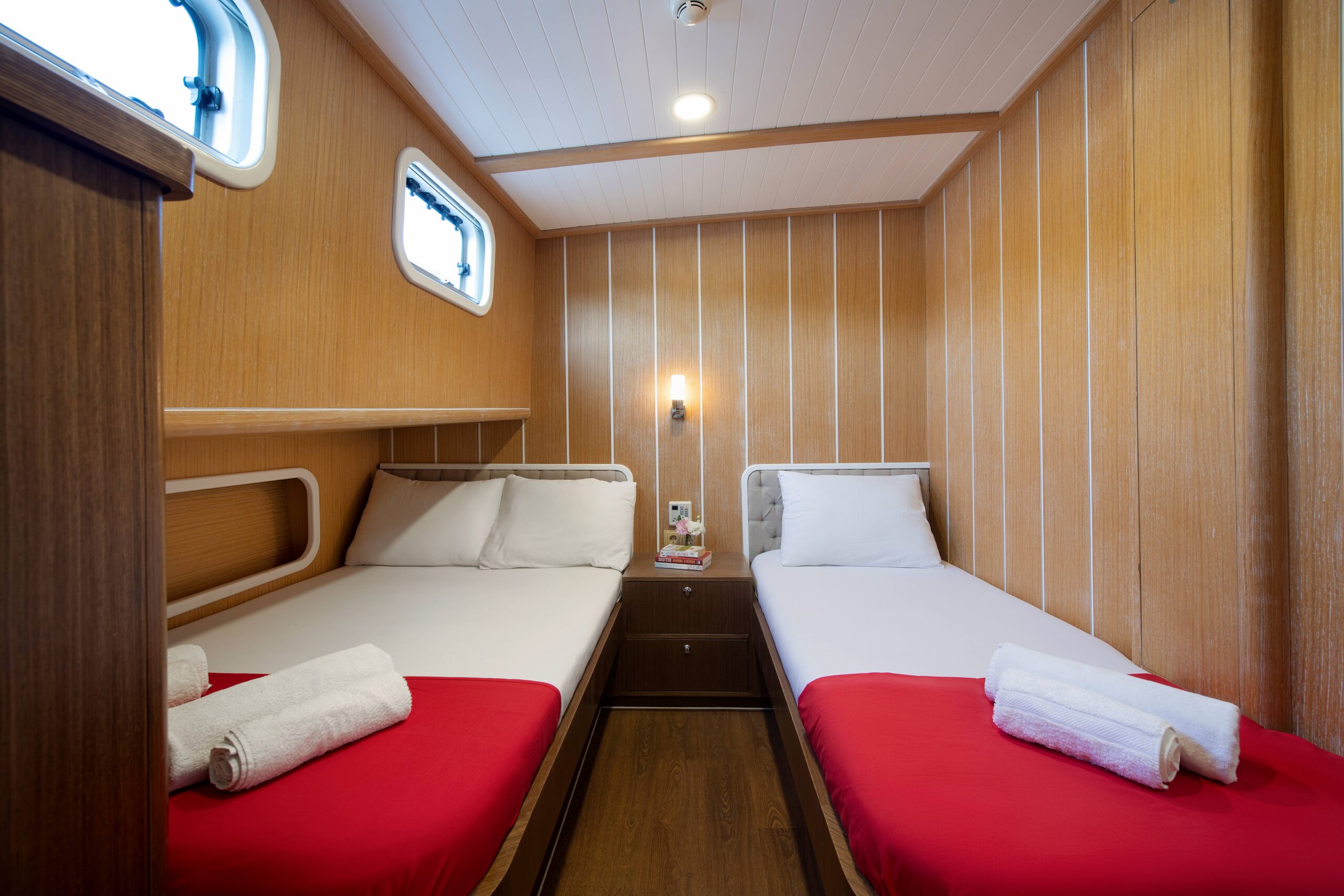 OGUZ BEY A cozy cabin with two single beds, each with white sheets and red blankets, neatly folded towels, a small nightstand with a magazine, wood-paneled walls, and two small windows.