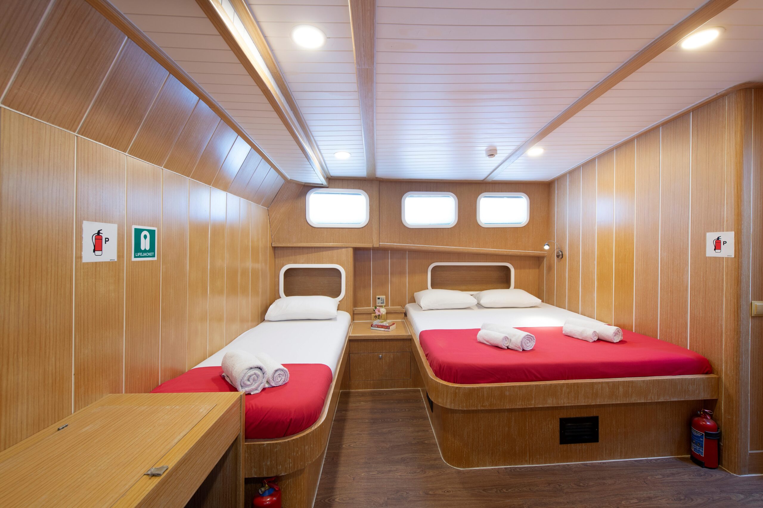 OGUZ BEY A cozy wooden cabin with two single beds, each with red bedding and rolled white towels. There are small windows above the beds and fire extinguishers on the floor. Safety signs are on the walls.