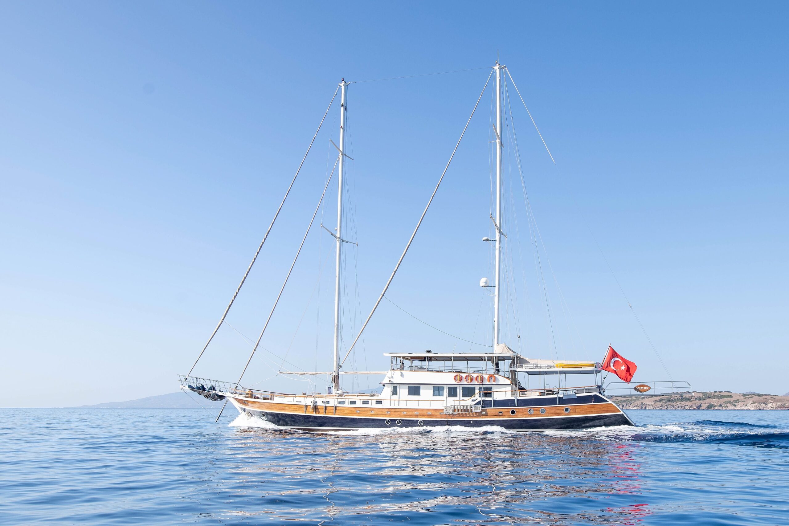OGUZ BEY A large wooden sailing yacht with two masts sails on calm blue water under a clear sky. A red Turkish flag is flying at the stern, and a distant coastline is visible in the background.