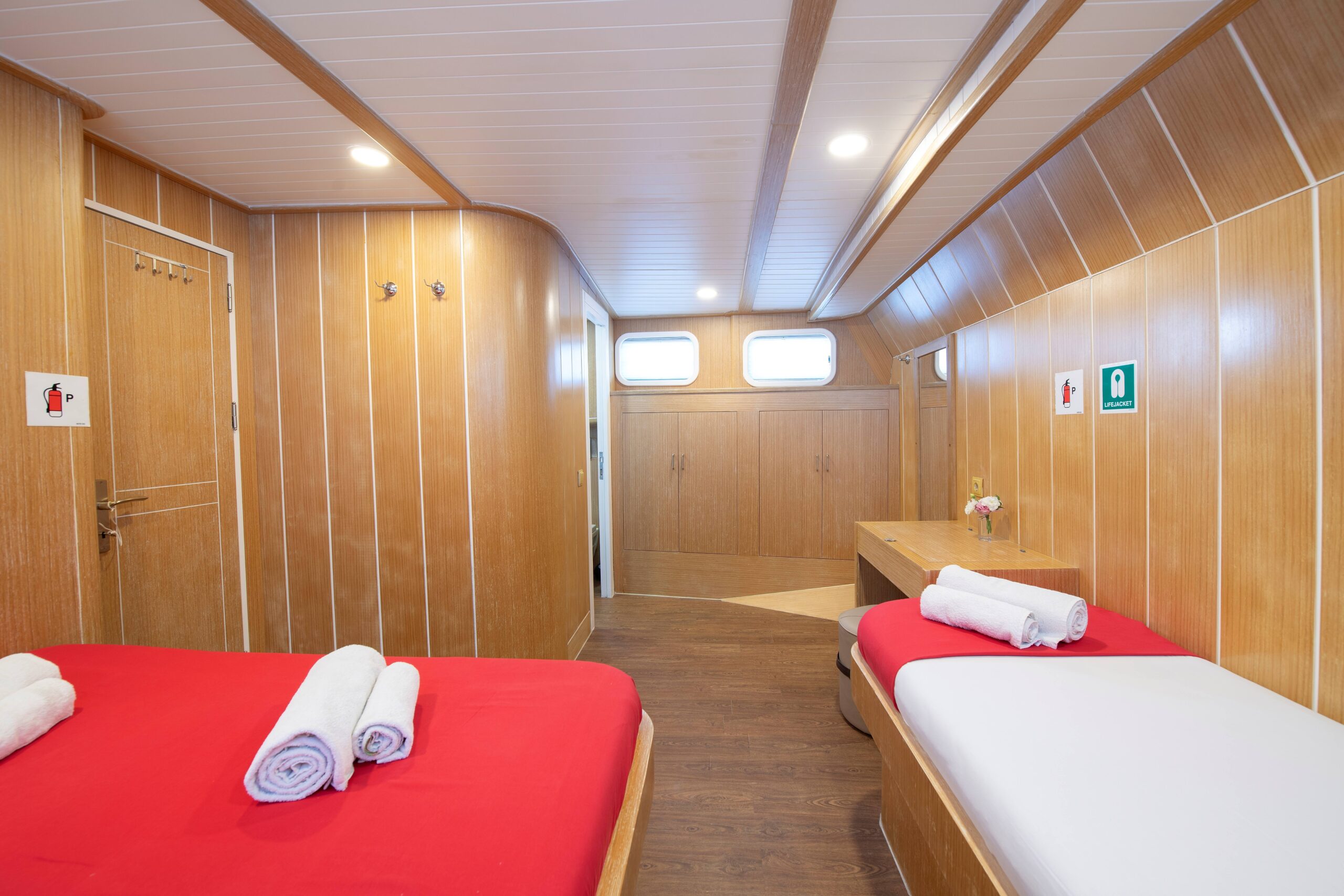 OGUZ BEY A cozy, wood-paneled room with two single beds dressed in red and white linens, each topped with rolled towels. The room has a desk with a small vase of flowers, wall hooks, and two rectangular windows near the ceiling.