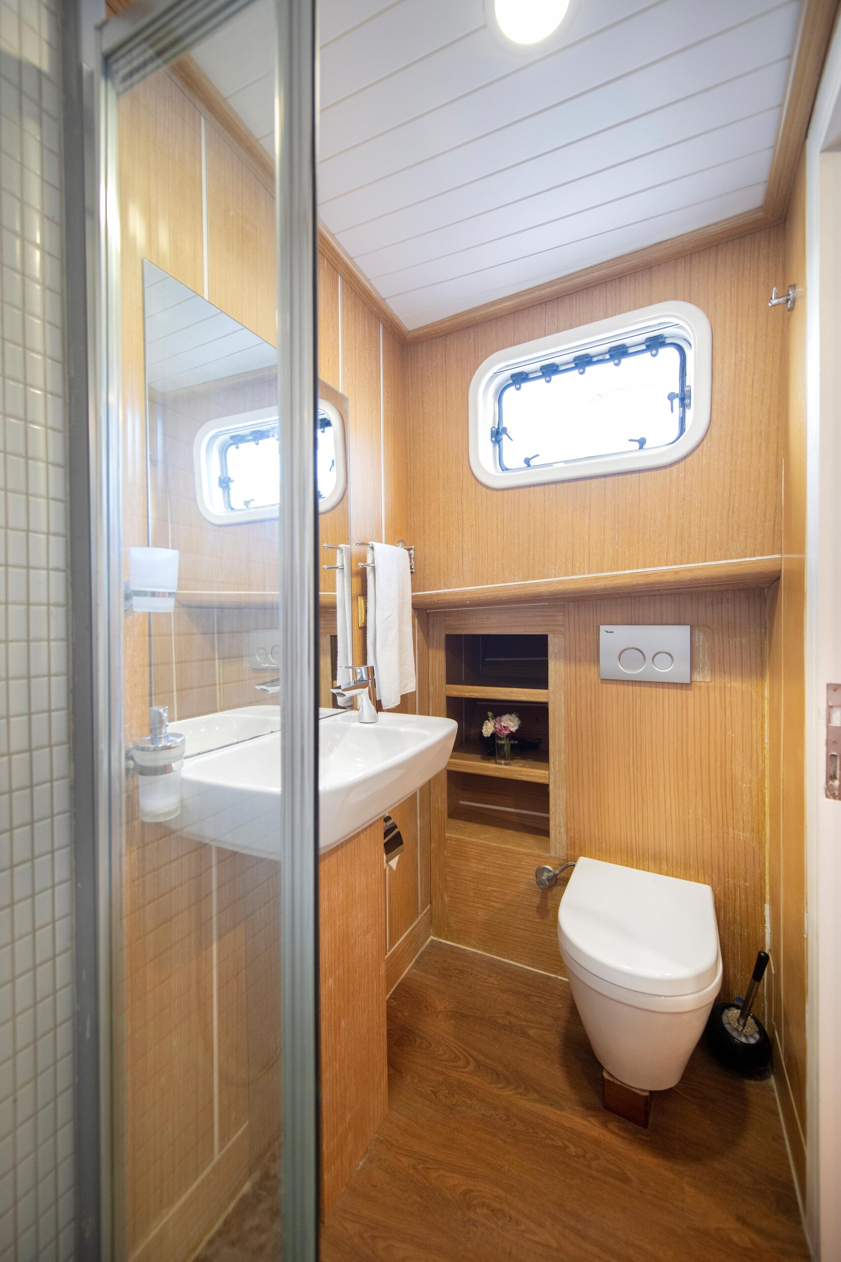 OGUZ BEY A compact bathroom with wood-paneled walls, a white sink with a mirror, a wall-mounted toilet, a small window, shelves with decor, and a shower area with a glass door.