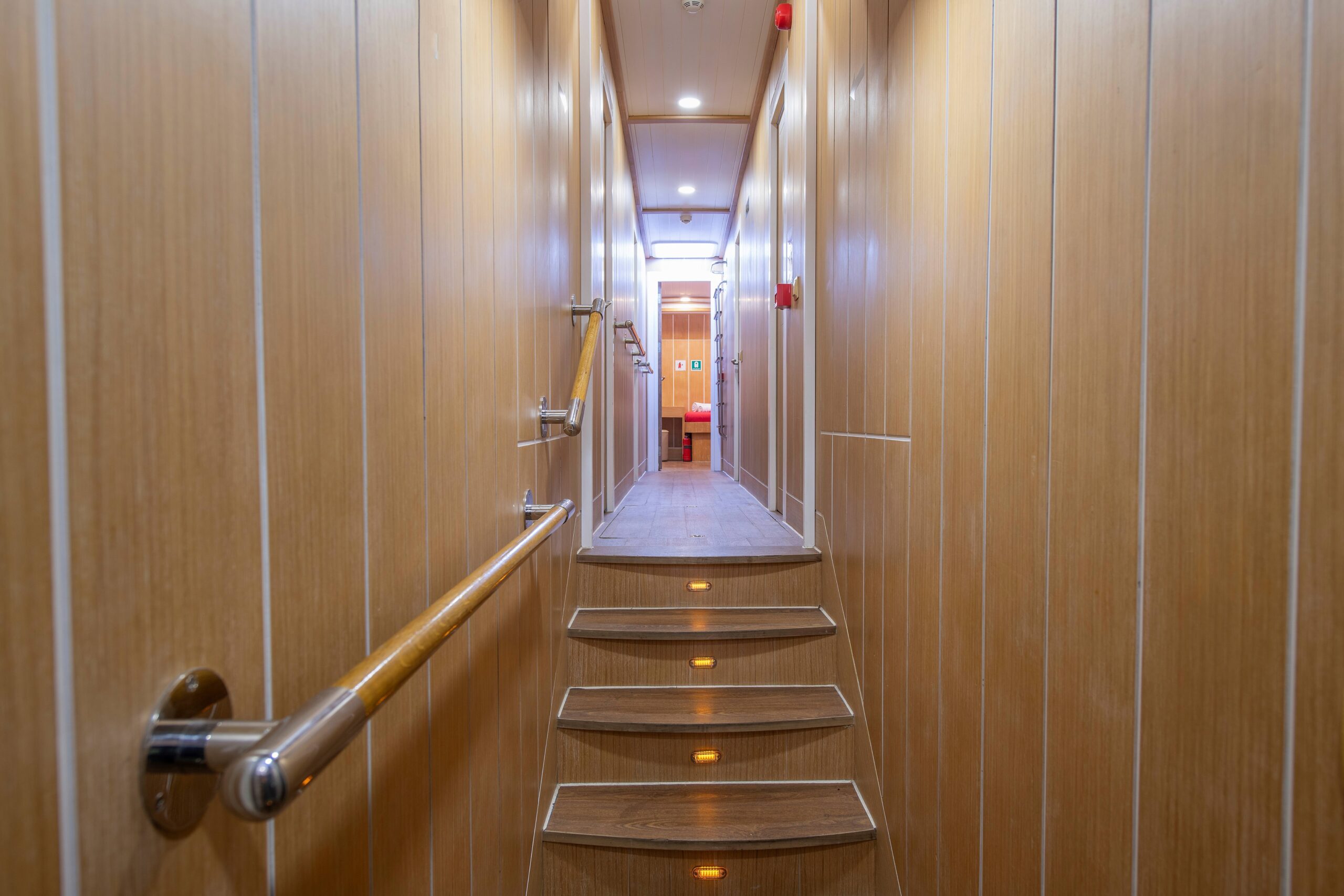 OGUZ BEY A narrow hallway with wooden walls and steps, handrails on the left, and lights along the steps, leading to a brightly lit area with a red object at the end.