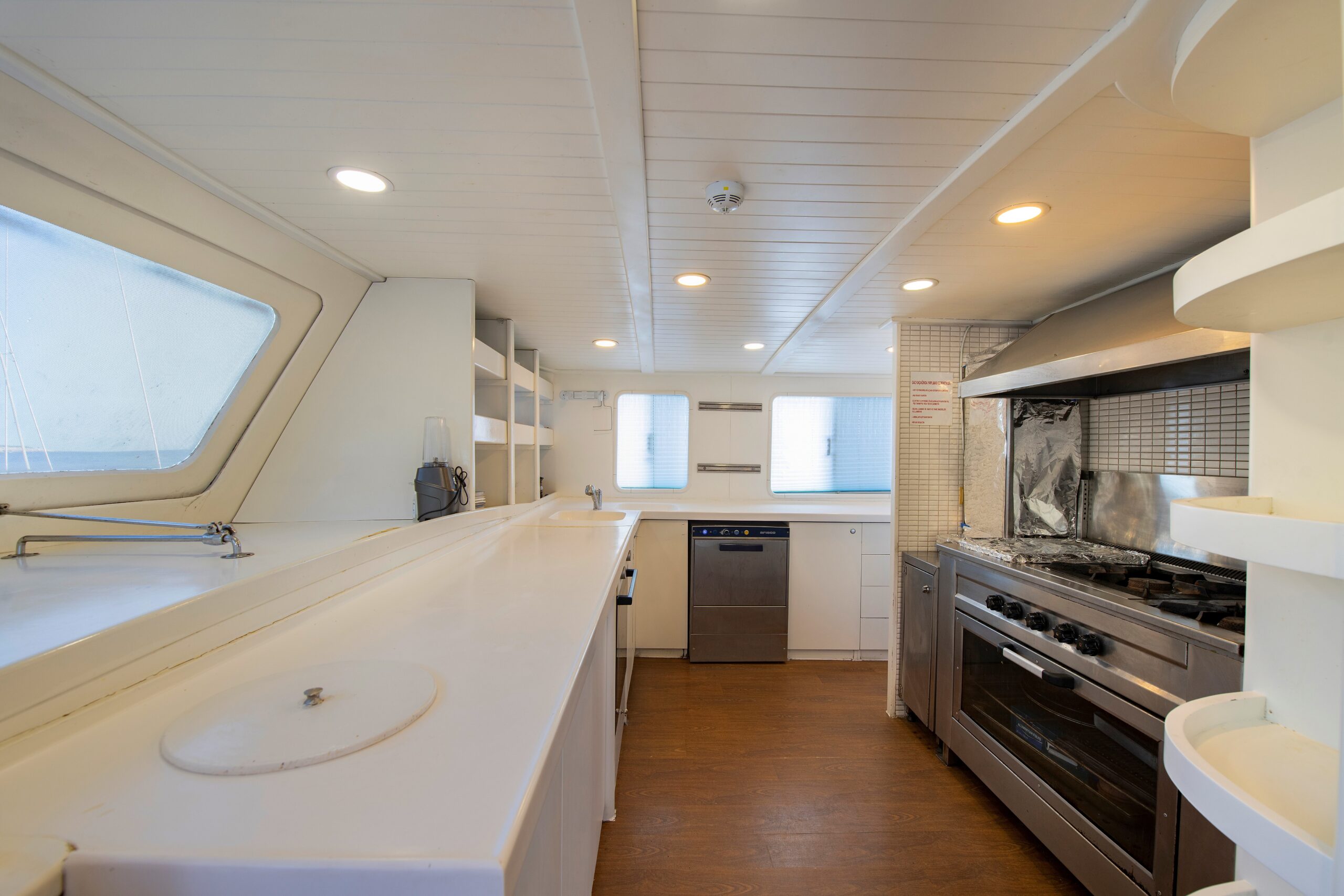 OGUZ BEY Spacious boat kitchen with large white countertops, stainless steel stove and oven, dishwasher, open shelving, wooden floor, and windows letting in natural light.