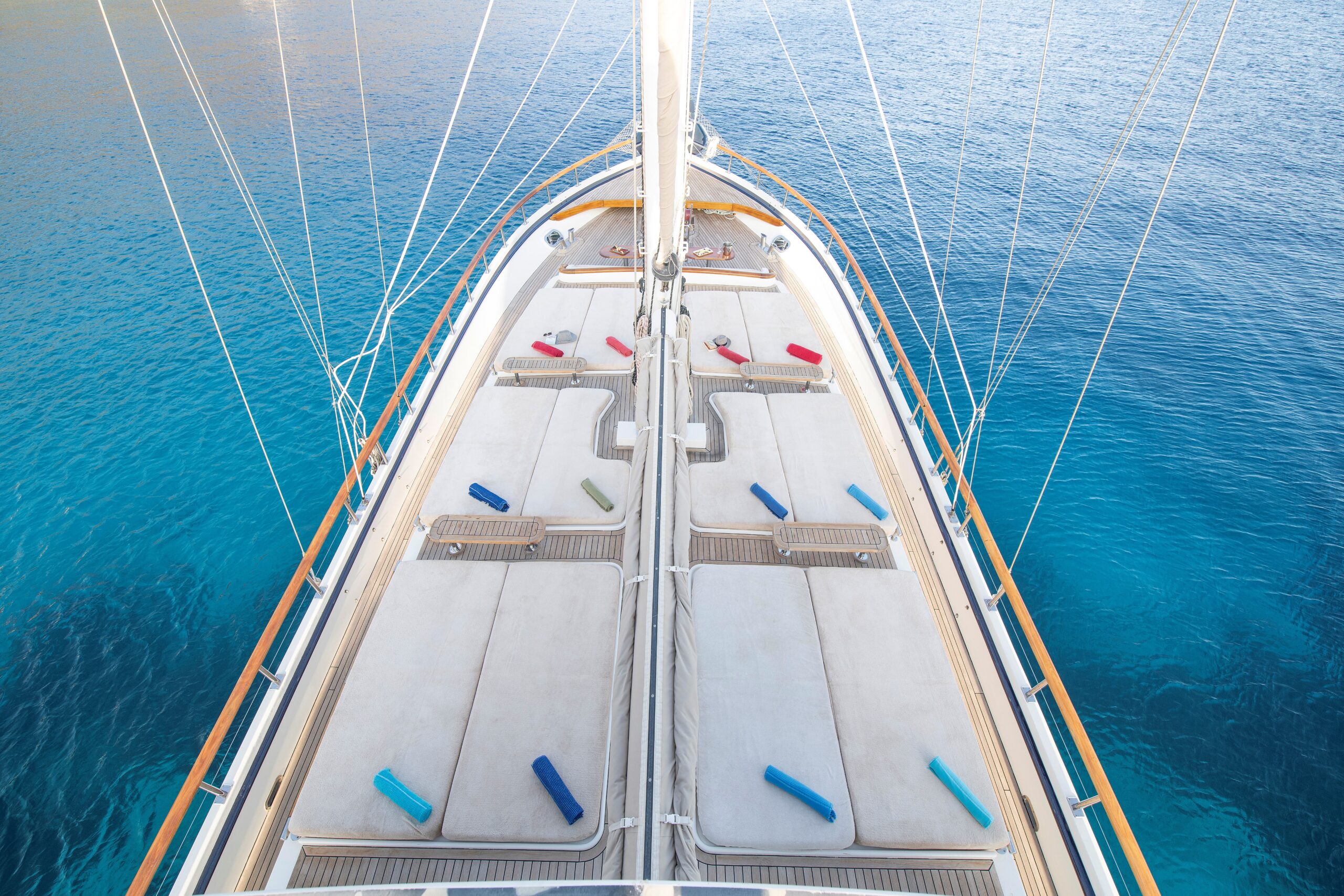 OGUZ BEY View from above of a yacht deck with white sunbeds and colorful cushions, surrounded by clear blue water. The yachts bow is visible, and the sea appears calm and inviting.