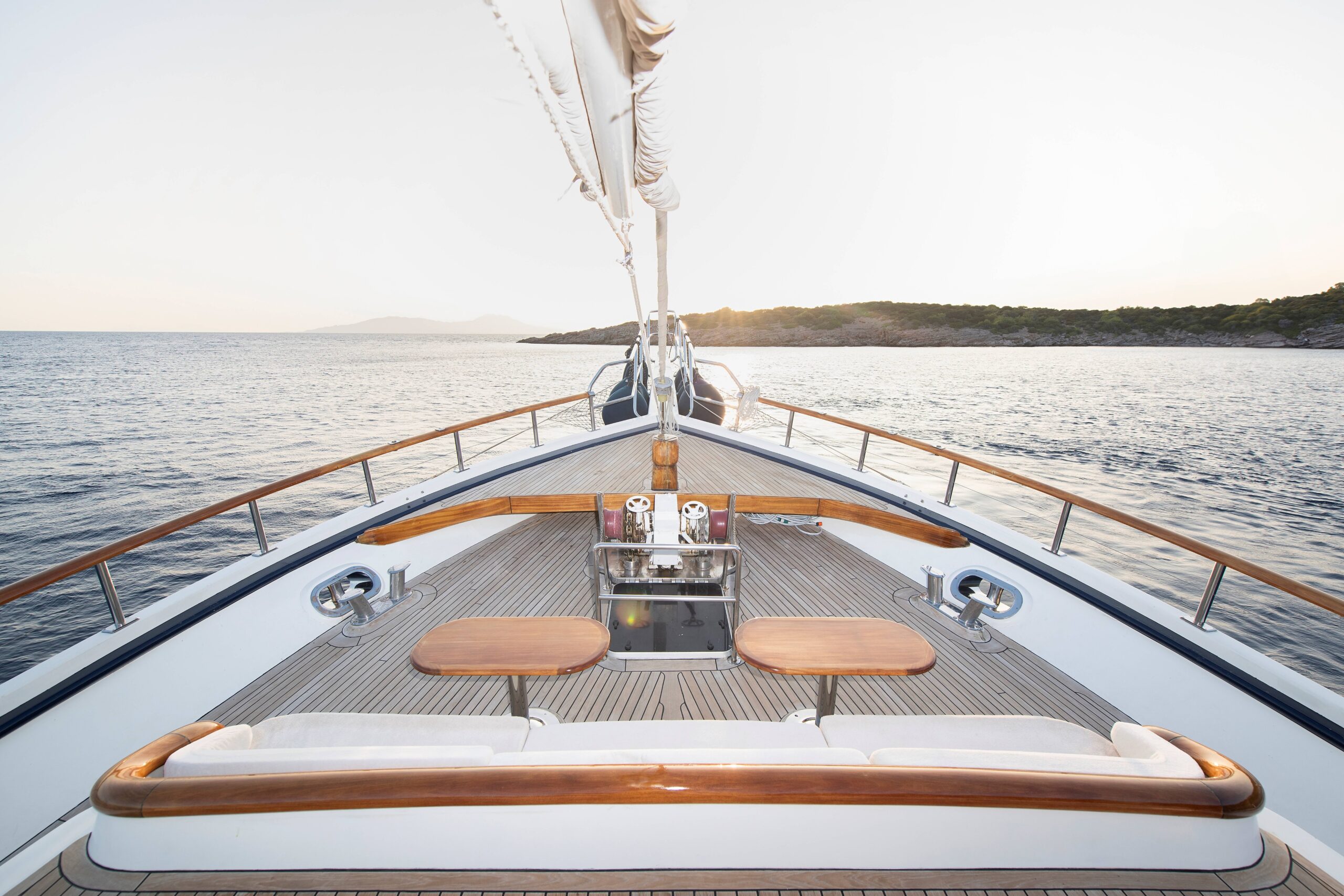 OGUZ BEY View from the deck of a yacht showing two wooden tables, a small sink area, and railing overlooking calm sea waters with a distant shoreline under a clear sky.