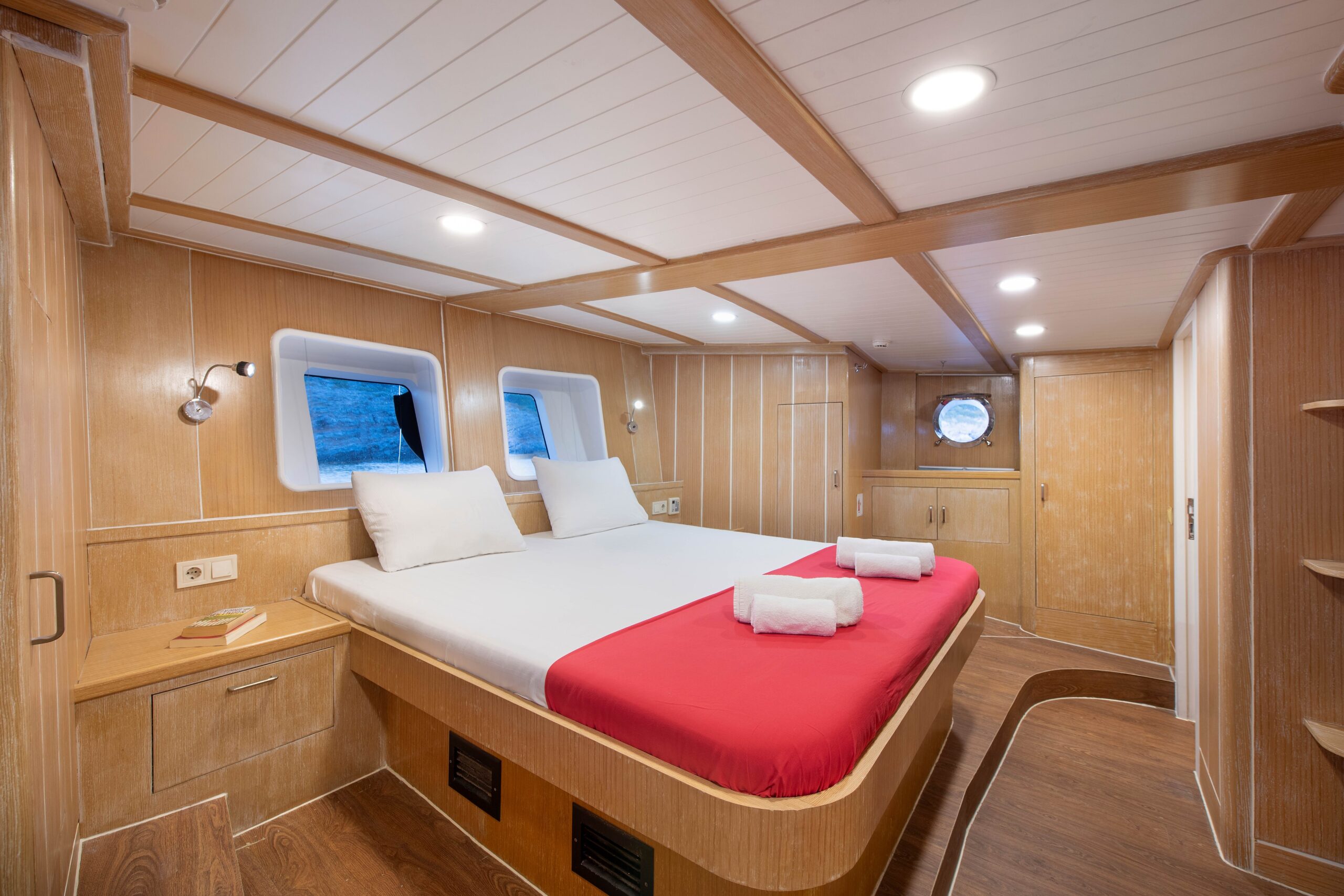 OGUZ BEY A cozy, modern ship cabin with a double bed covered in white linens and a red blanket, two rolled towels, wood-paneled walls, built-in shelves, windows, and soft overhead lighting.