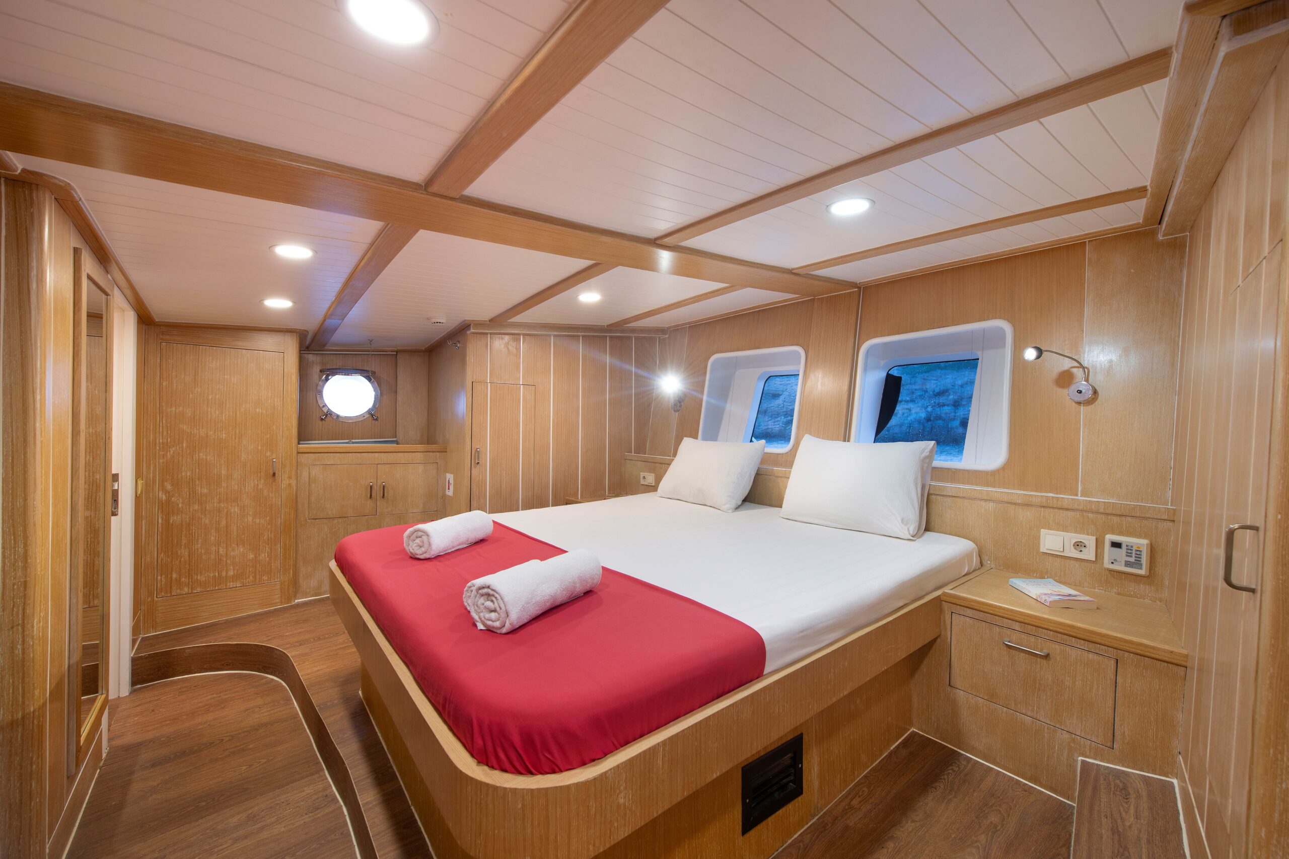 OGUZ BEY A cozy, well-lit cabin with wood-paneled walls, a large bed with red sheets and white pillows, two rolled towels, small bedside tables, and two windows showing an ocean view.