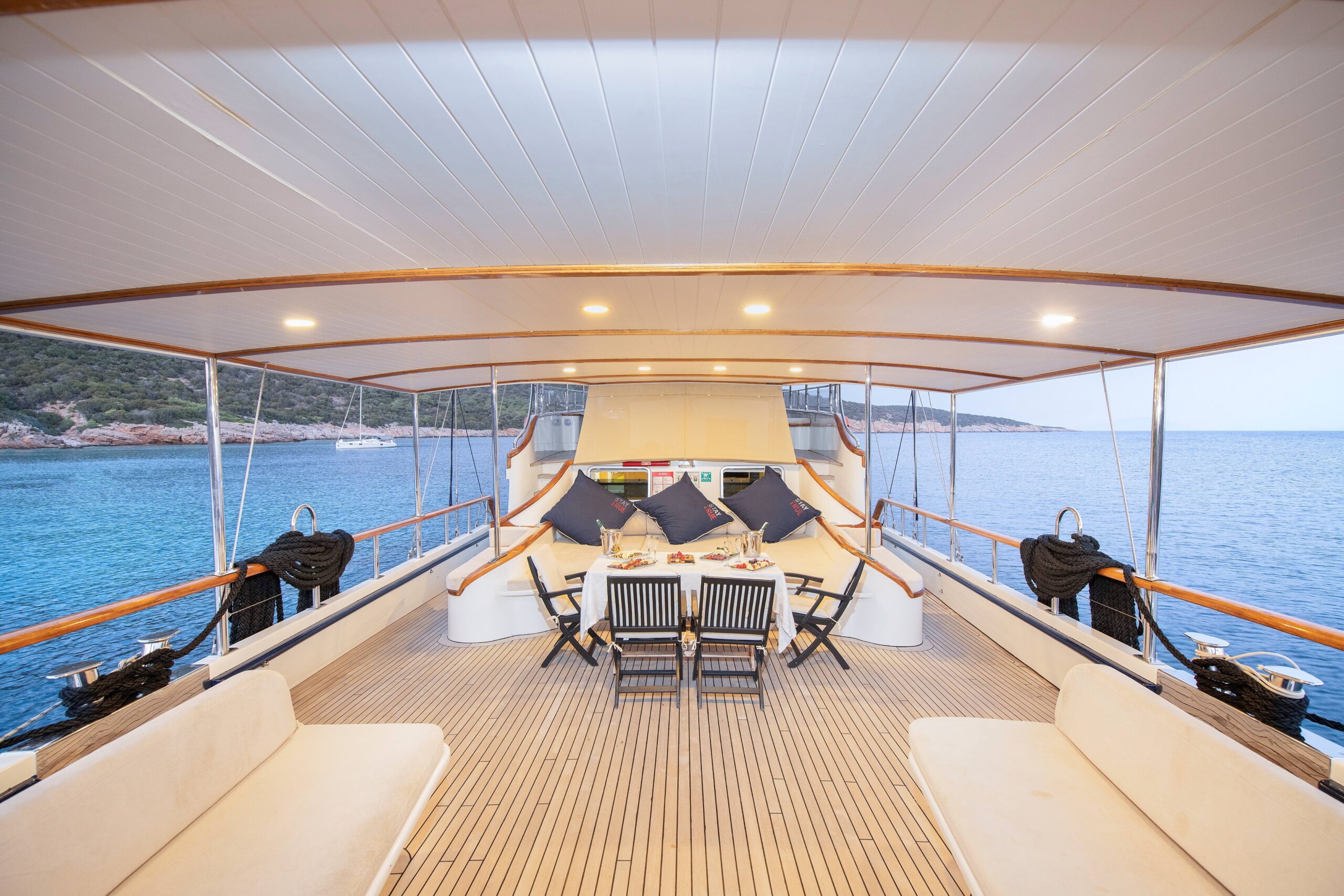 OGUZ BEY A spacious yacht deck with wooden flooring features a dining table set for six, surrounded by cushioned benches. The yacht is anchored near a rocky shoreline with calm, blue water in the background.