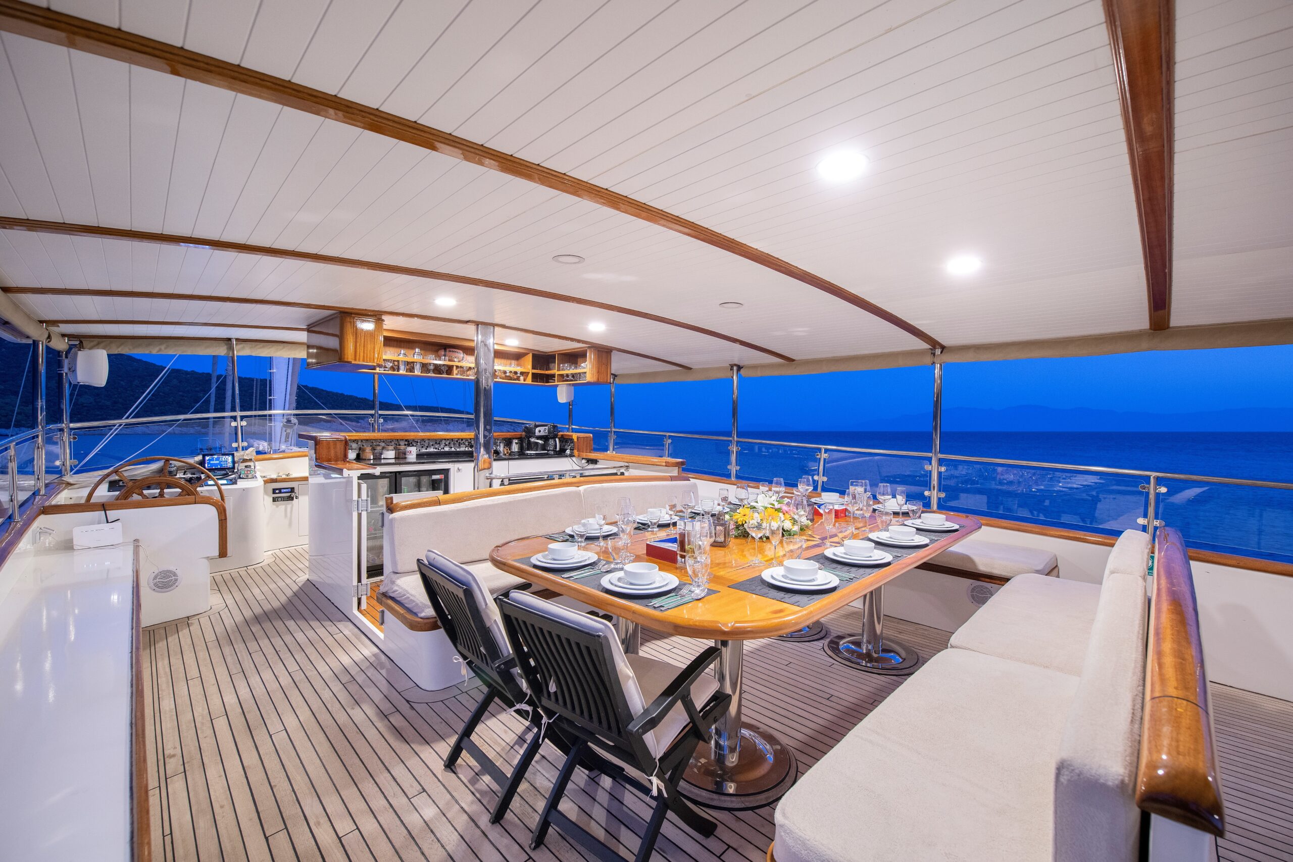 OGUZ BEY A luxurious yacht deck set for dinner with a wooden table, cushioned seating, plates, glasses, and flowers, surrounded by open sea and mountains at dusk.