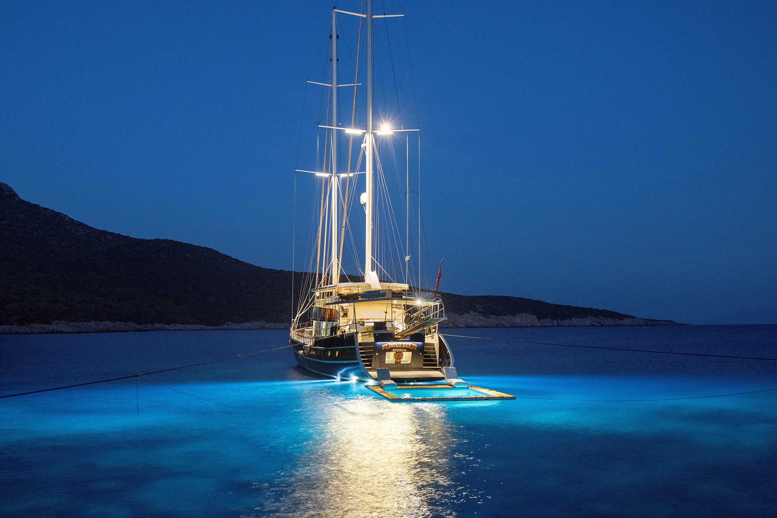 OGUZ BEY A large sailboat is anchored at night in calm blue waters, illuminated with bright underwater lights that create a glowing area around the boat. Hills and a dark sky are visible in the background.