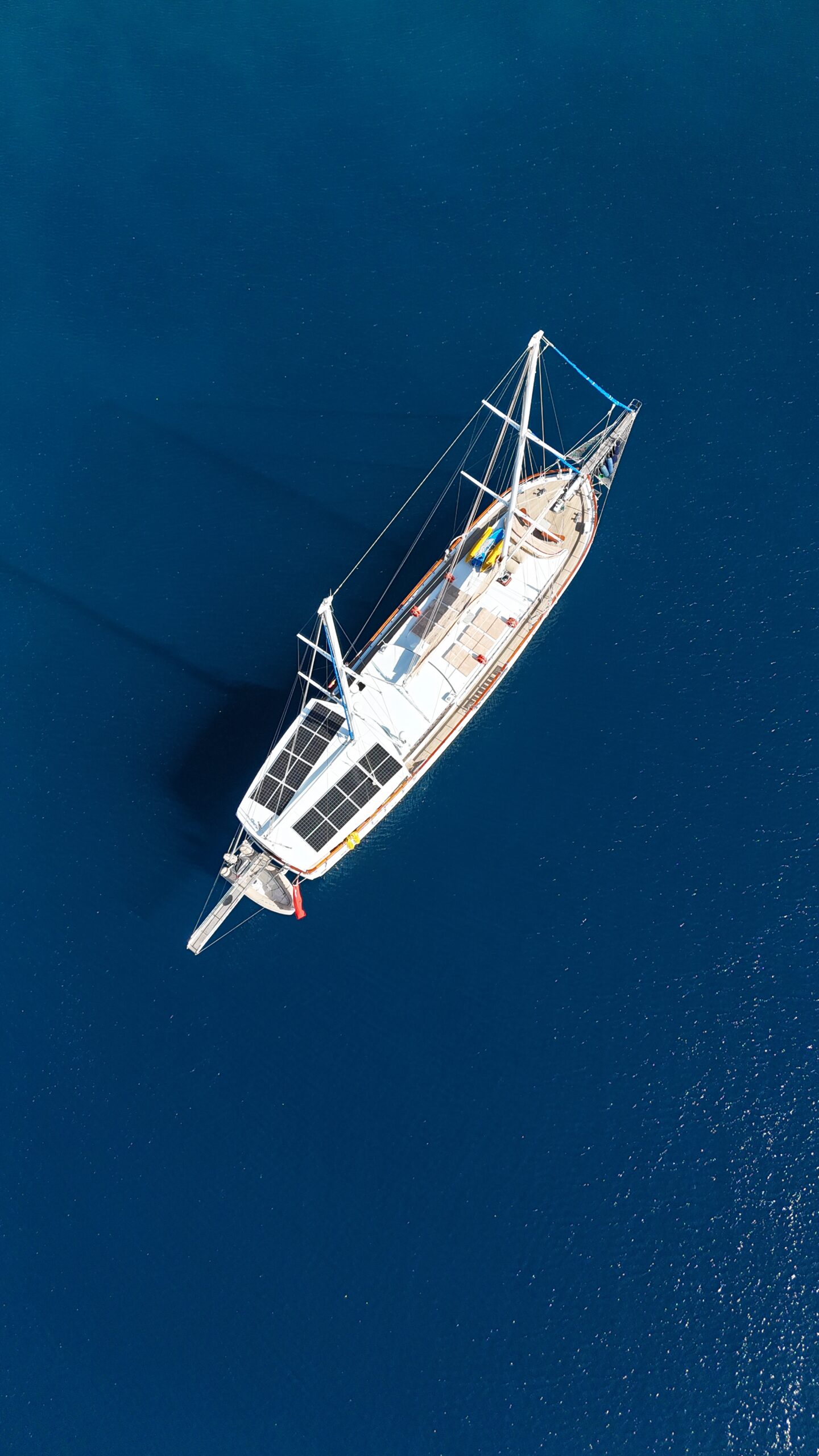 CEMRE 4 A white sailboat with two masts floats on calm, deep blue water, viewed from above. The boat casts a shadow on the water, and no other boats or land are visible in the scene.
