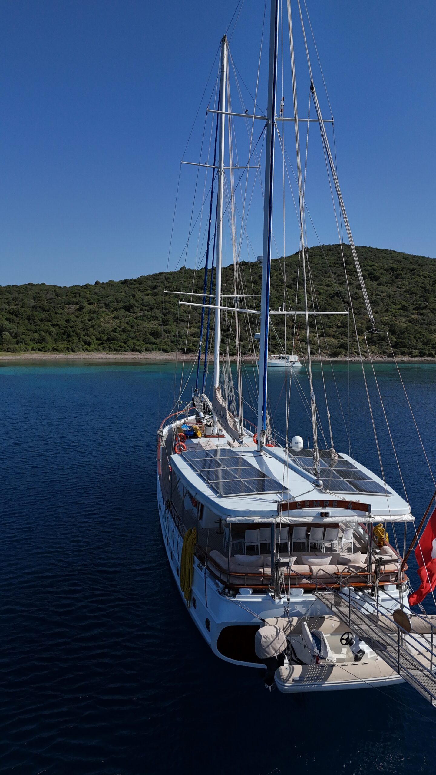 CEMRE 4 A white sailboat with solar panels is anchored in calm blue water near a lush, green coastline under a clear blue sky.