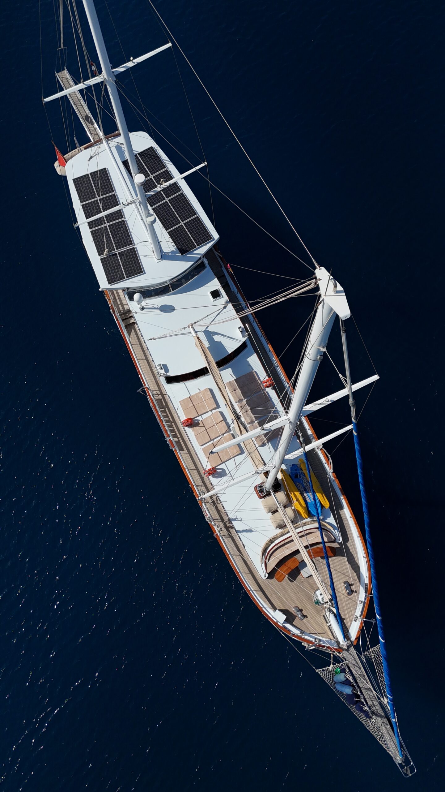 CEMRE 4 Aerial view of a large sailboat with solar panels on the roof, wooden decking, and two masts, floating on calm, deep blue water.